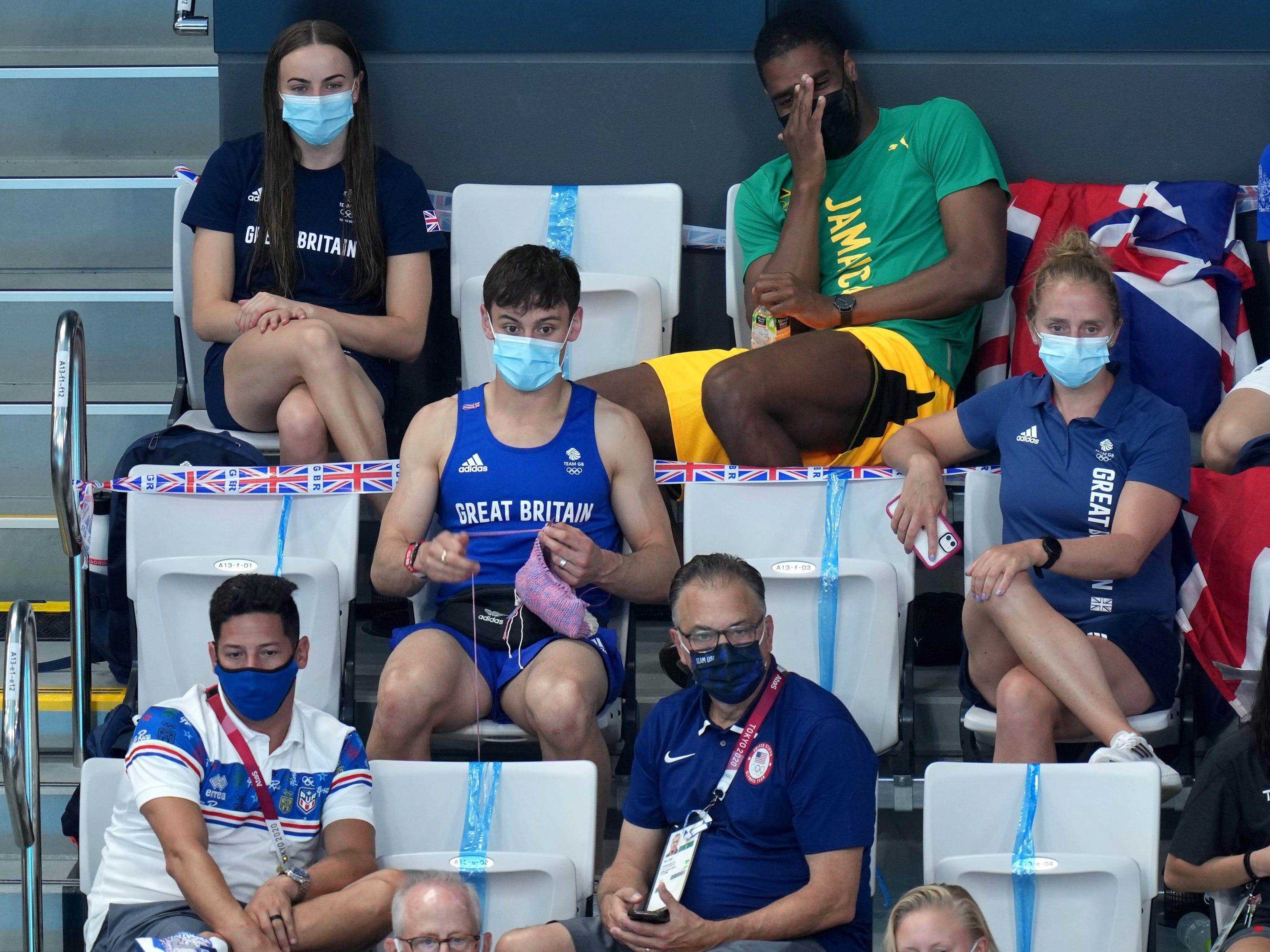 Tom Daley sitzt auf der Zuschauertribüne bei den Olympischen Spielen in Tokio und strickt inmitten des Sprungbrettspringens der Frauen.