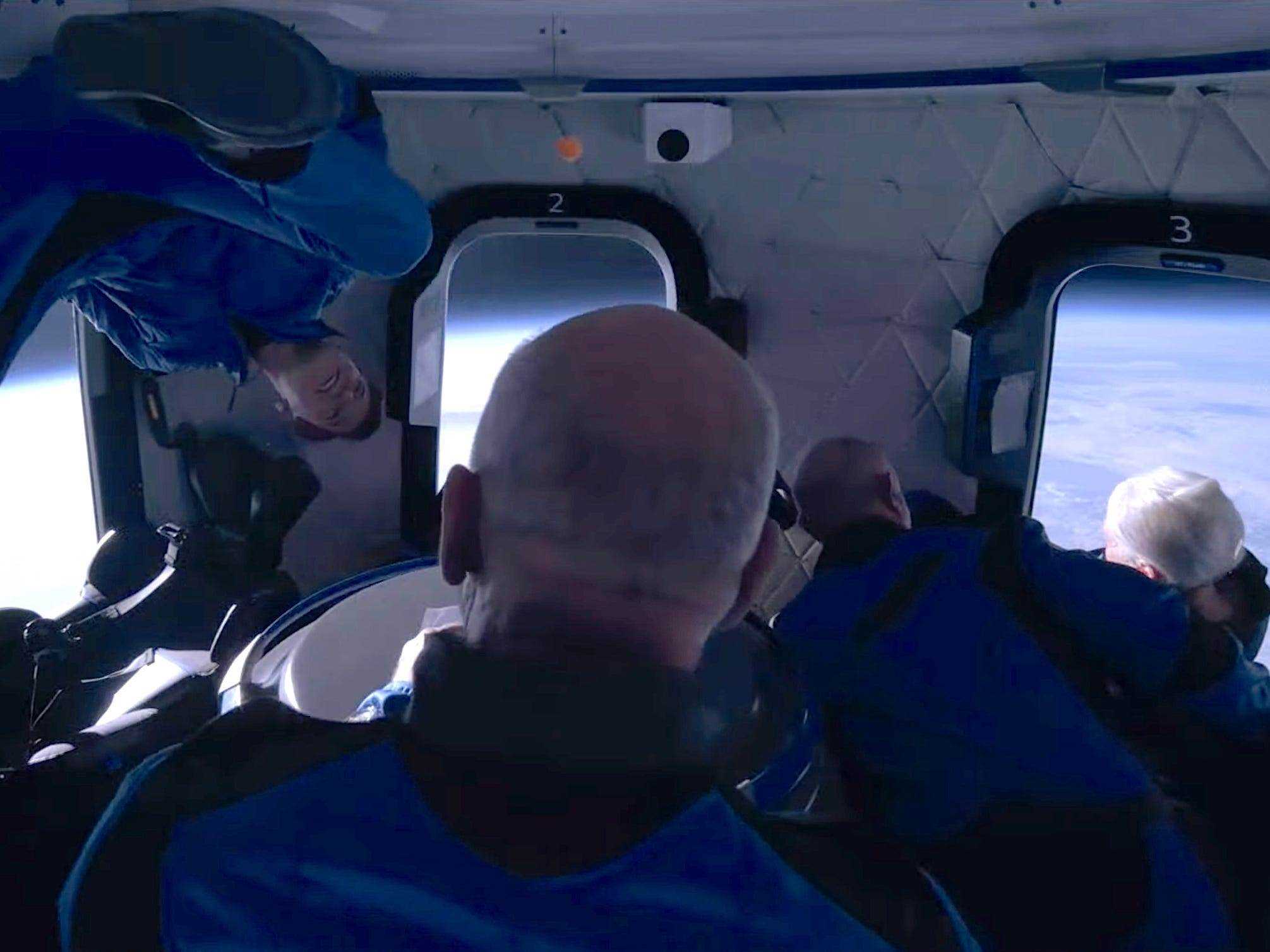 Jeff Bezos und drei andere Passagiere in Overalls schweben im Hintergrund um die Erde der Raumschiffkabine herum