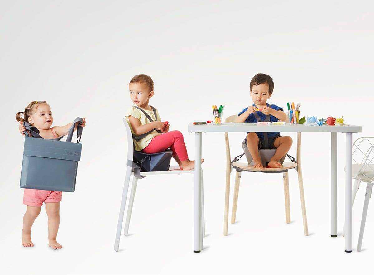 zwei Kleinkinder, die in Kinderstühlen am Tisch sitzen und ein Kleinkind, das einen zusammengeklappten Kinderstuhl trägt