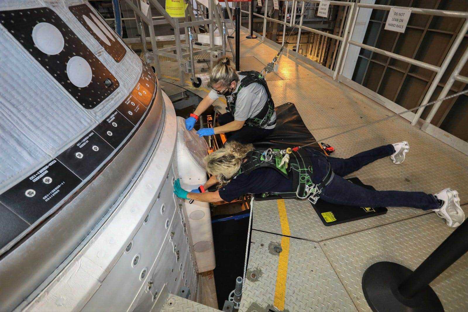 Zwei Ingenieure, die Gurte mit Bungee-Leinen tragen, arbeiten an einem Starliner-Raumschiff auf einer Rakete