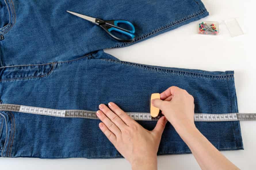 Kaukasische Frauenhände messen die gewünschte Länge mit einem Maßband auf einem weißen Tisch.  Kürzen Sie die Jeans mit Schere und Nähnadel.  DIY Sommerkleidung.  Ansicht von oben.
