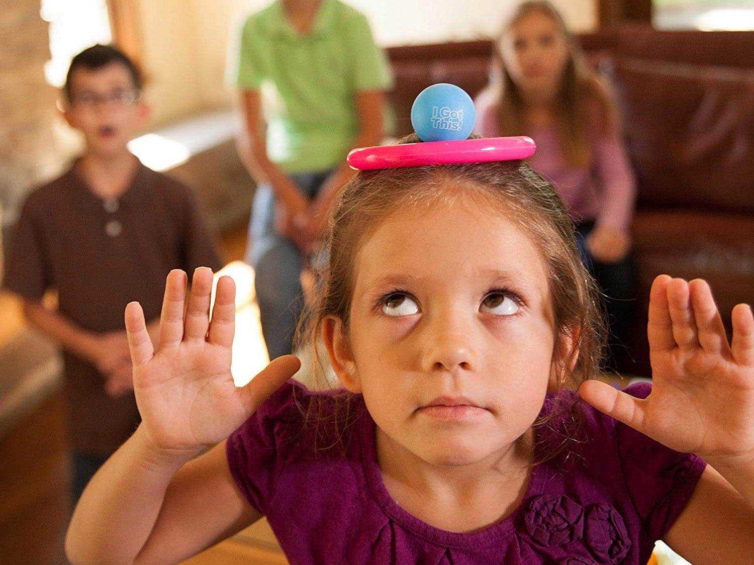 Mädchen balanciert einen Ring und einen Ball auf ihrem Kopf, während die Familie zusieht, während sie I Got This Spiel spielt