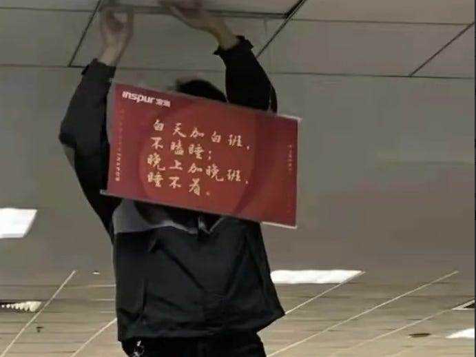 Mann, der ein Banner in chinesischer Sprache aufstellt, in dem Überstunden gepriesen werden.