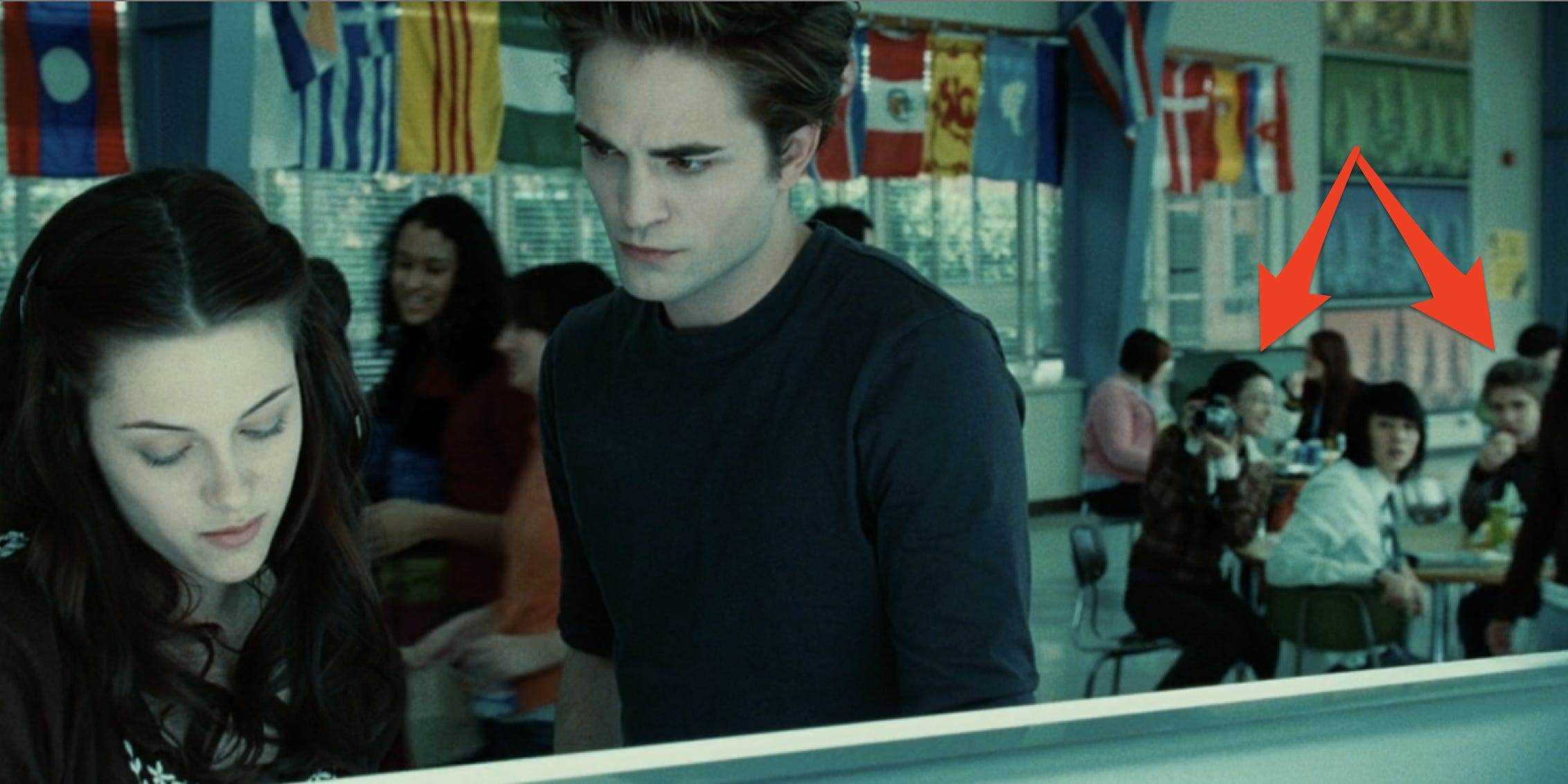 Bella und Edward unterhalten sich in der Mittagsschlange mit drei ihrer Freunde, die im Hintergrund an einem Tisch sitzen und sie anstarren