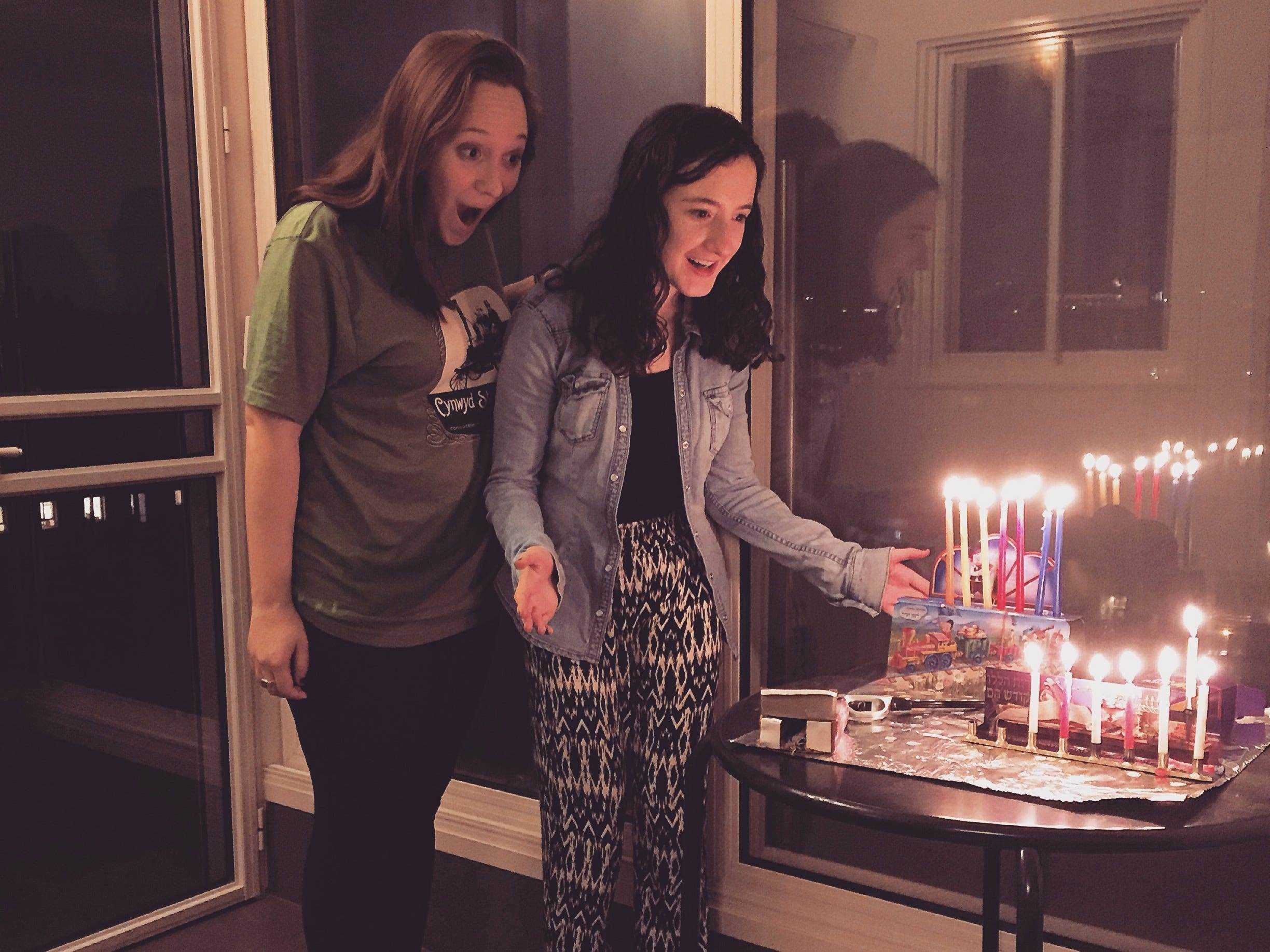 Zwei Frauen betrachten eine mit Chanukka-Kerzen beleuchtete Menora mit glücklichem Ausdruck.