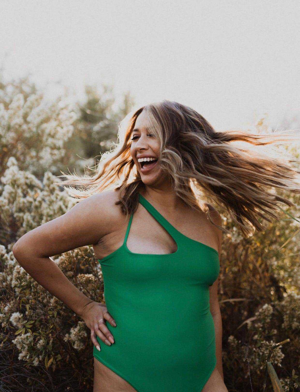 Frau mit grünem Arrow + Phoenix-Badeanzug lacht im Freien mit Hand auf Hüfte