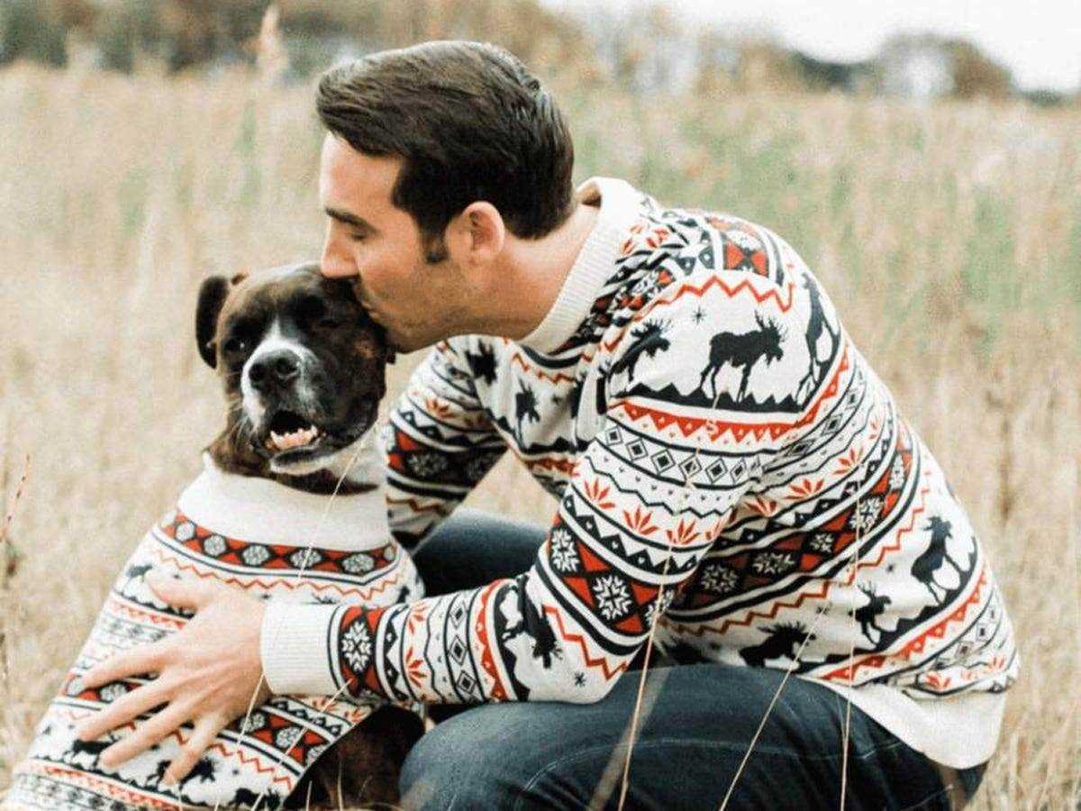 Eine Person, die einen Pullover mit Weihnachtsmuster trägt und einen schwarzen Hund küsst, der denselben bedruckten Pullover trägt