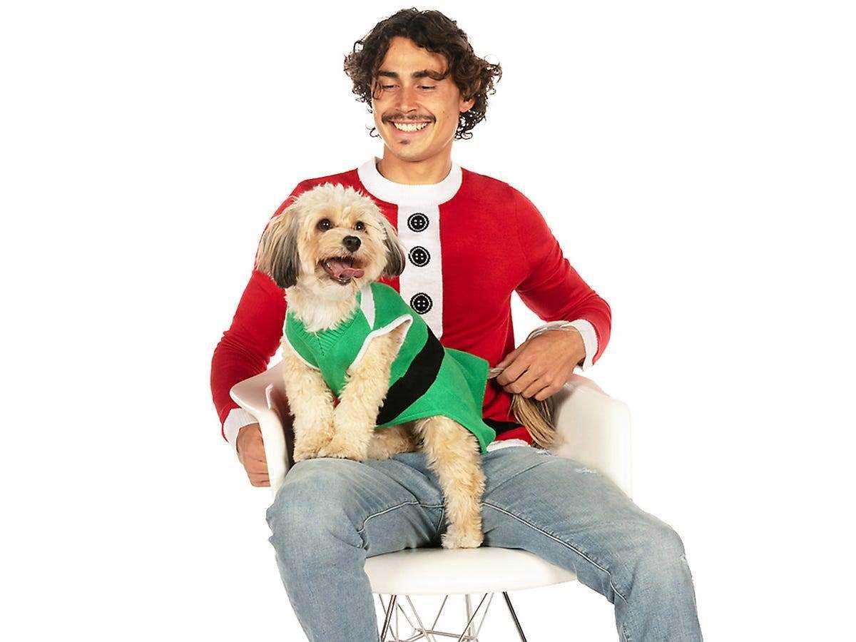 Ein lächelnder hellbrauner Hund, der einen grünen Pullover trägt und auf einem Menschen sitzt, der einen roten Weihnachtspullover trägt