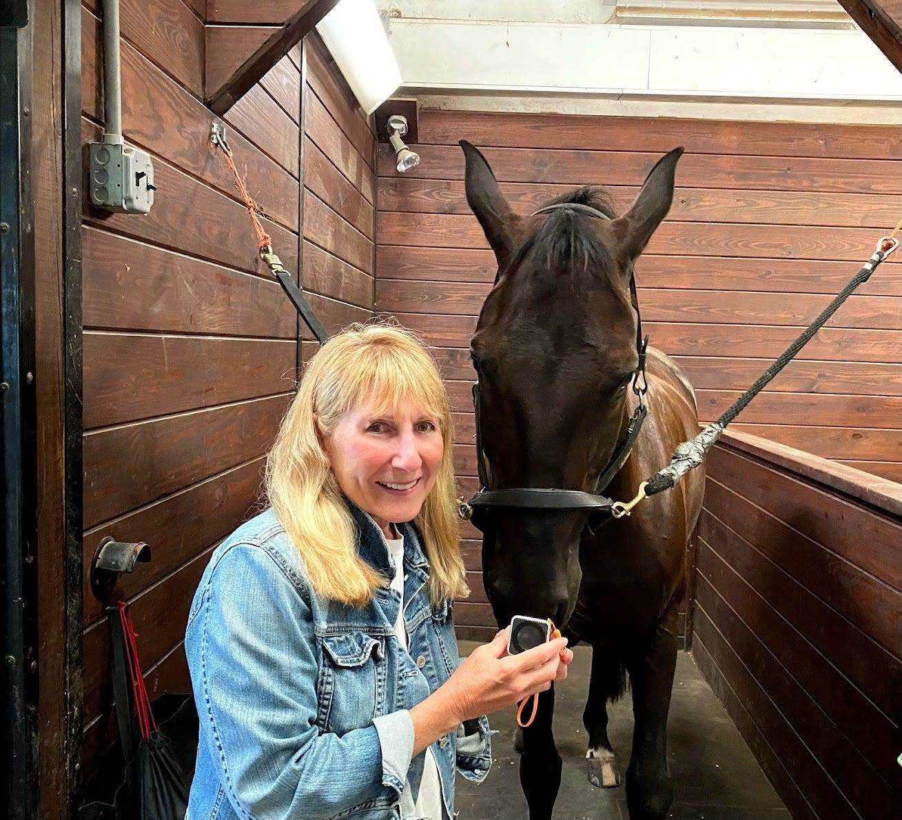 Janet Marlow, die blonde Haare und eine Jeansjacke trägt, lächelt neben einem Pferd in einem Stall.