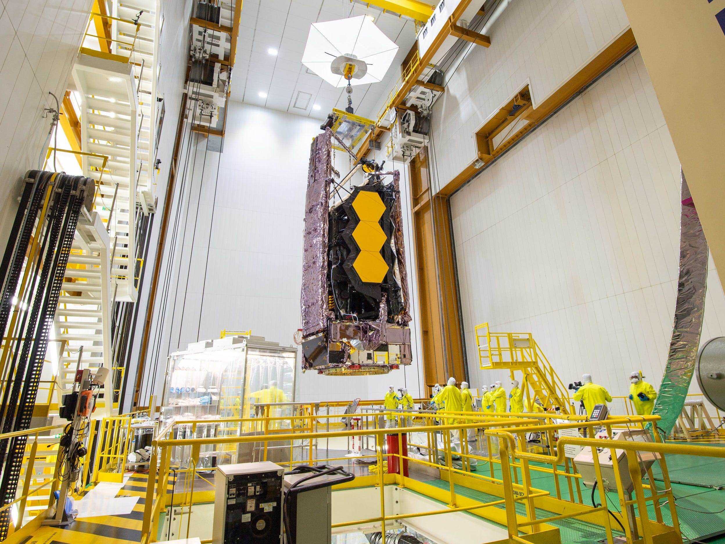 James Webb Weltraumteleskop zusammengeklappt hängend über Loch mit gelbem Geländer