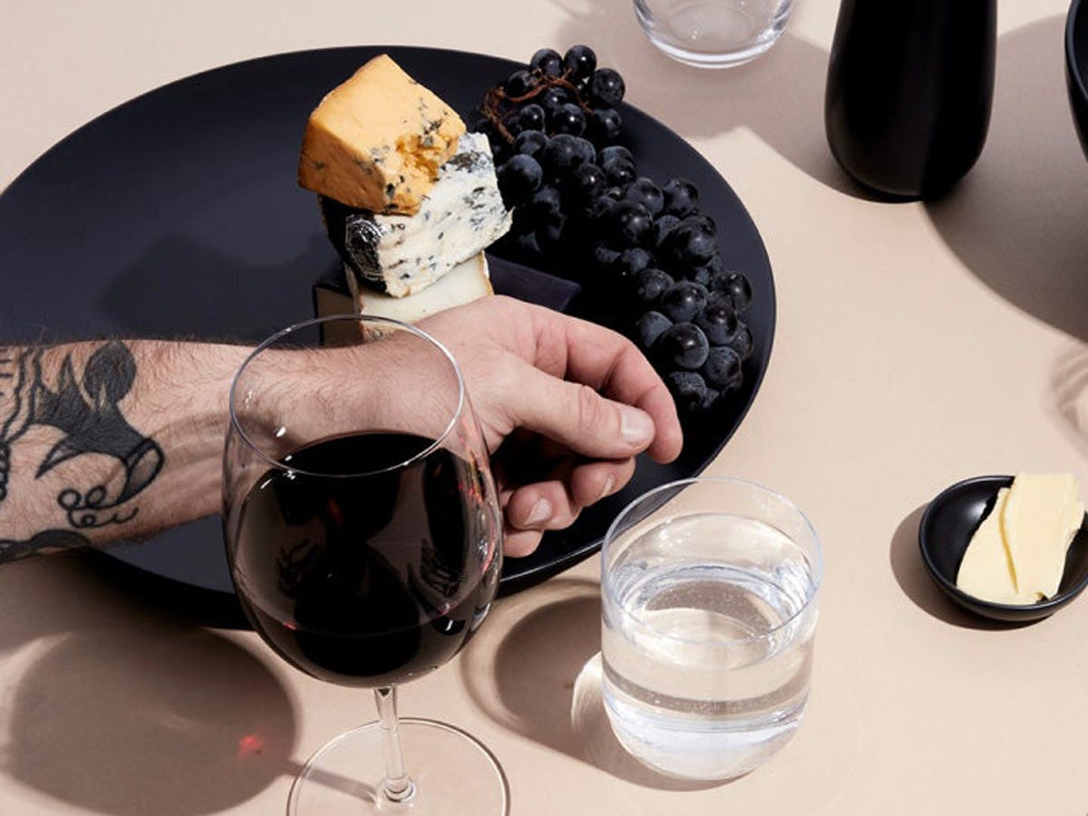 eine Hand ruht auf dem Rand eines schwarzen Tellers mit Trauben und Käse, ein Glas Rotwein zur Seite