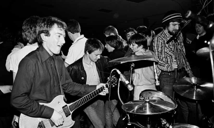 Schwarz-Weiß-Foto eines jungen Andy Gill, der neben dem Schlagzeug Gitarre spielt, ganz in der Nähe von tanzenden Mitgliedern des Publikums hinter ihm