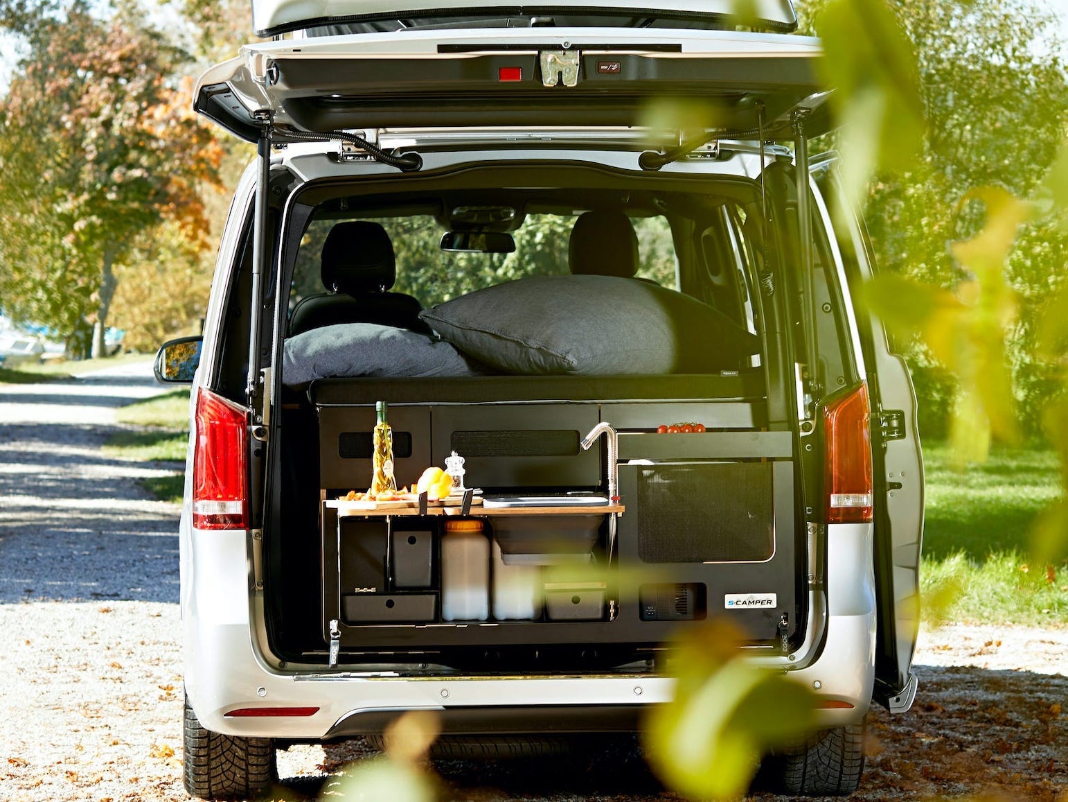 Der offene Kofferraum des EQV gibt den Blick auf eine Miniküche frei.  Der Van ist zwischen Bäumen geparkt.