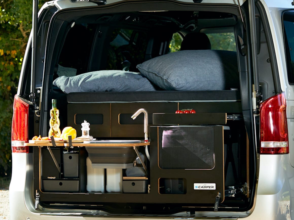 Der Kofferraum des EQV lässt sich öffnen und gibt den Blick auf eine Miniküche frei.  Der Lieferwagen ist neben einem Essplatz im Freien geparkt.