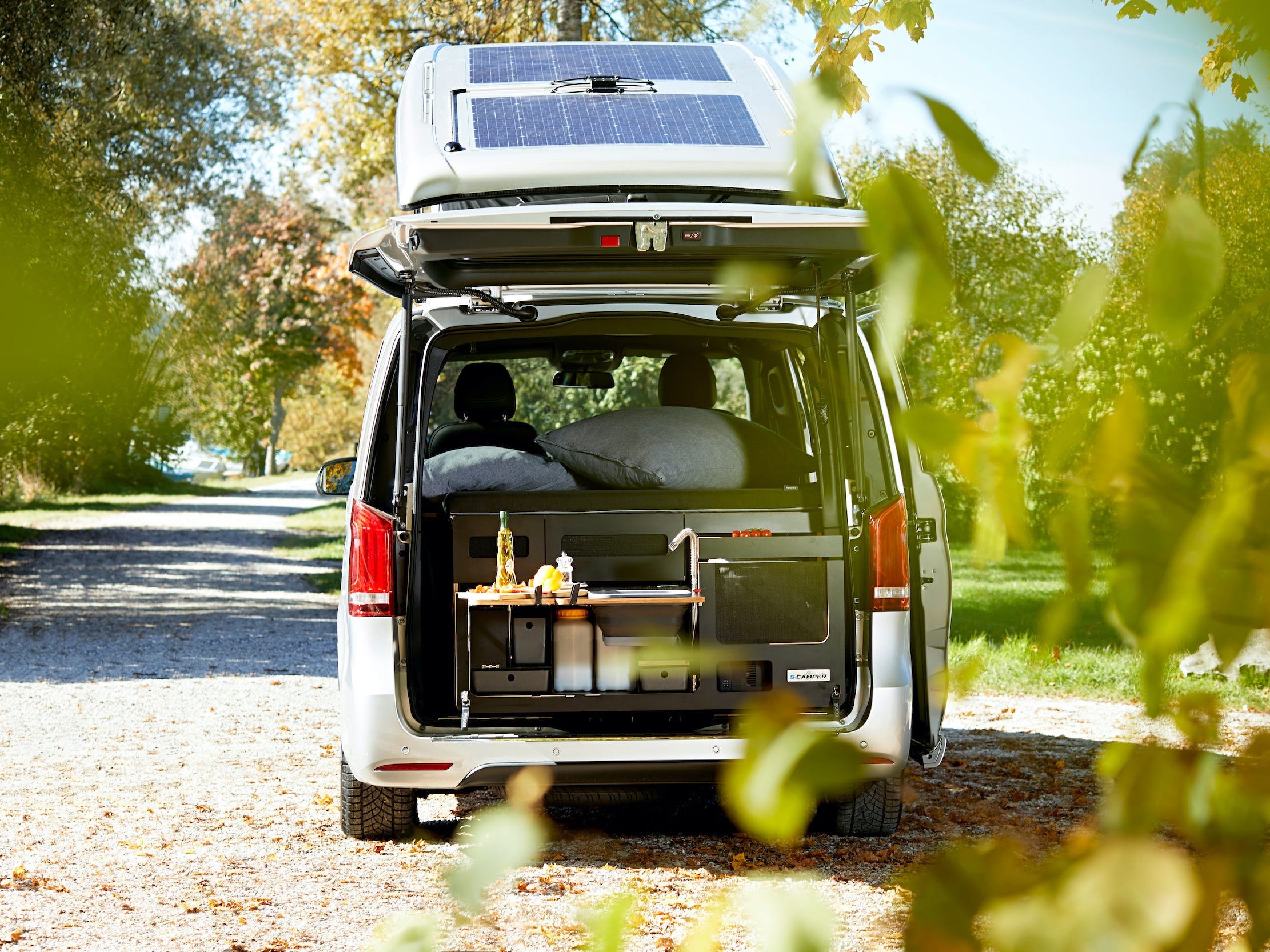 Der offene Kofferraum des EQV gibt den Blick auf eine Miniküche frei.  Der Van ist zwischen Bäumen geparkt.