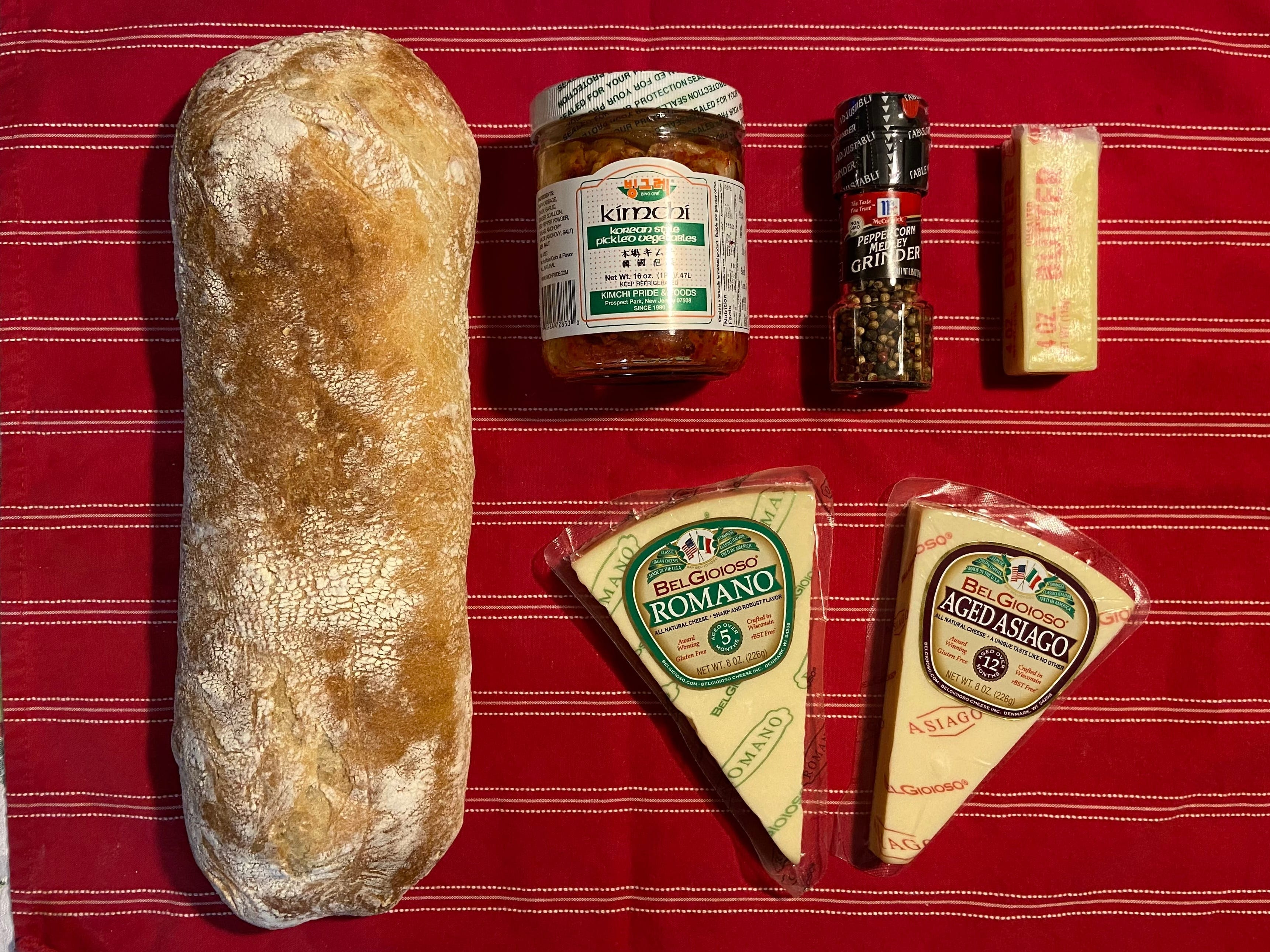 Zutaten auf einem Geschirrtuch, darunter eine Ladung Ciabatta-Brot, Romano- und Asiago-Käse, eine Flasche Kimchi, Pfefferkörner und Butter.