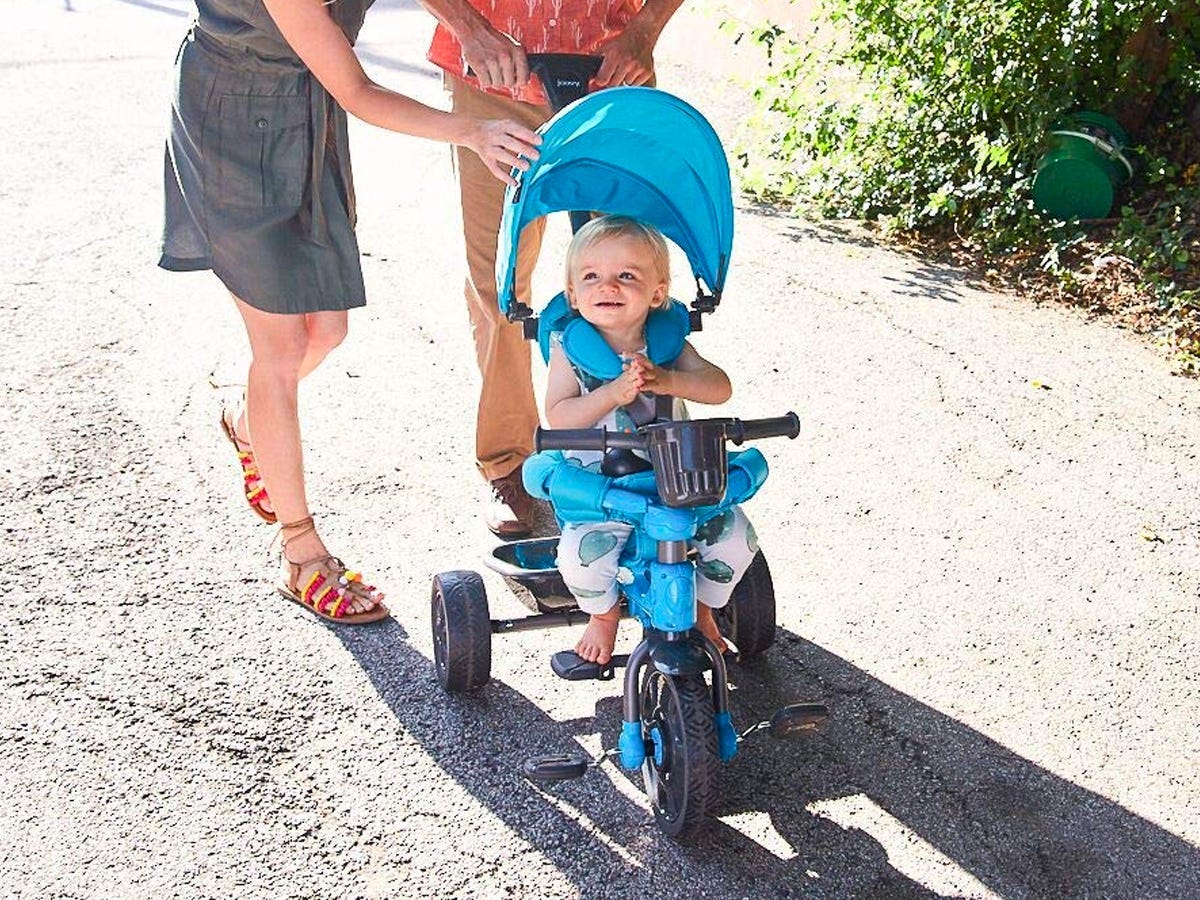 Zwei lächelnde Erwachsene, die hinter einer blauen Kombination aus Dreirad und Kinderwagen schleifen, auf der ein Kind sitzt und lächelt