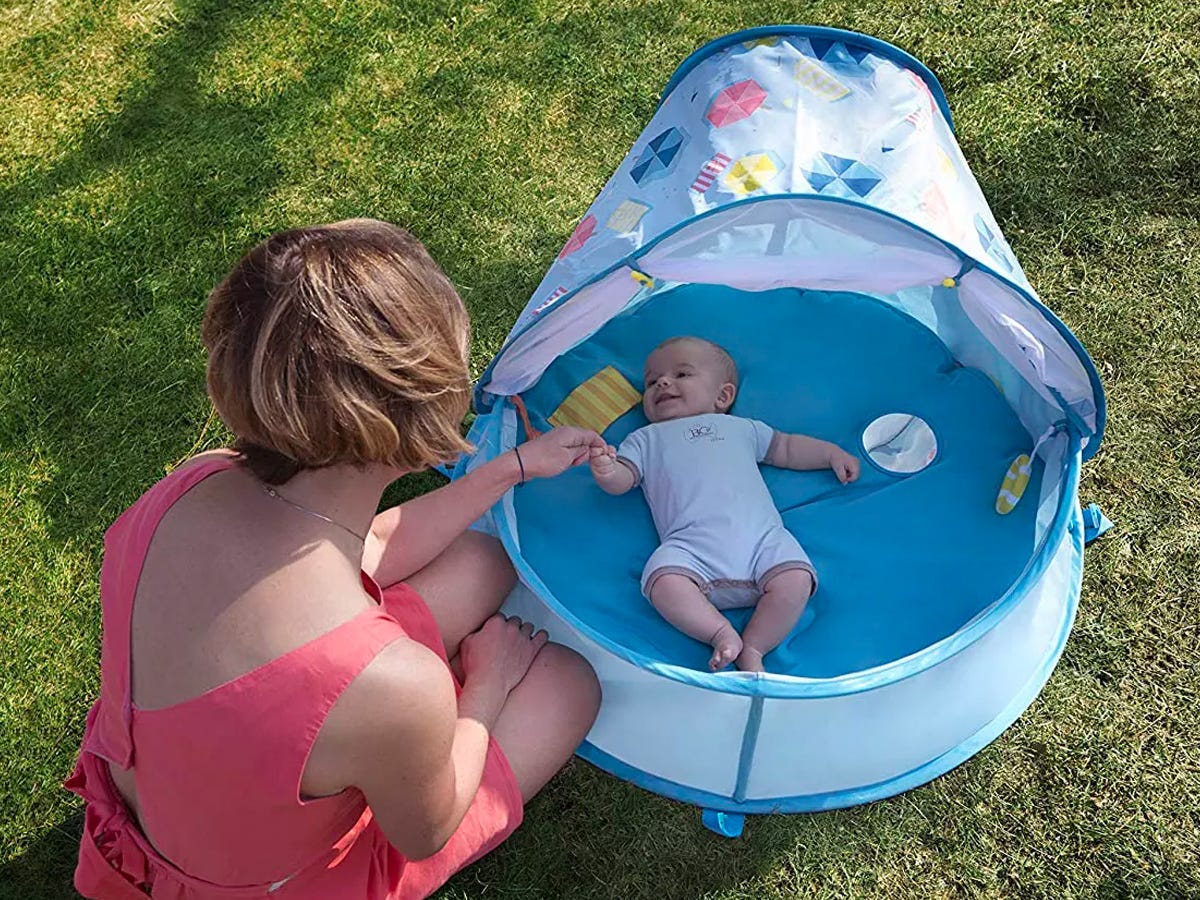 Baby liegt in einem blauen runden Spielplatz im Gras und ein Erwachsener sitzt auf dem Boden neben dem Greifen, um die Hand des Babys zu halten