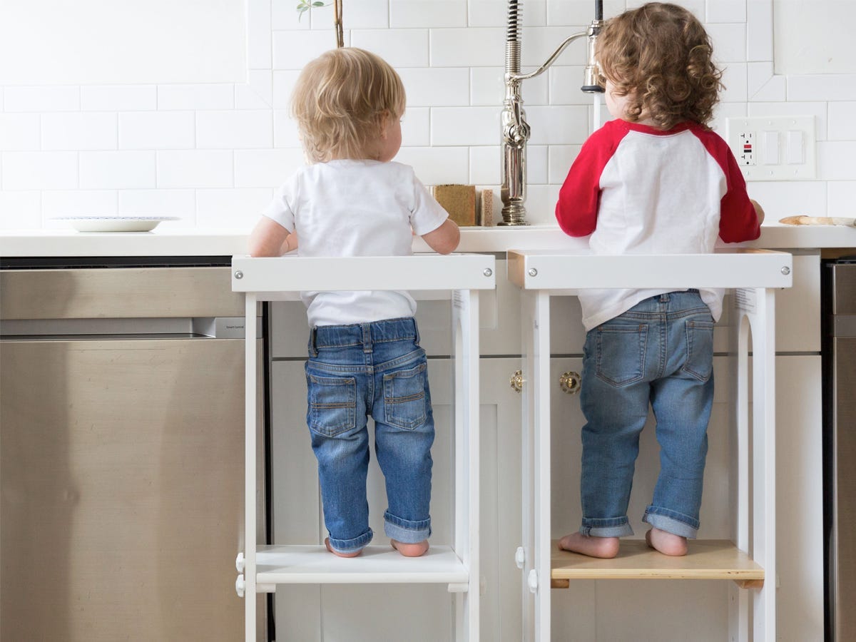 Zwei Trittleitern vor einer Küchenspüle, in der einen steht ein jüngeres Kleinkind, in der anderen ein älteres Kleinkind