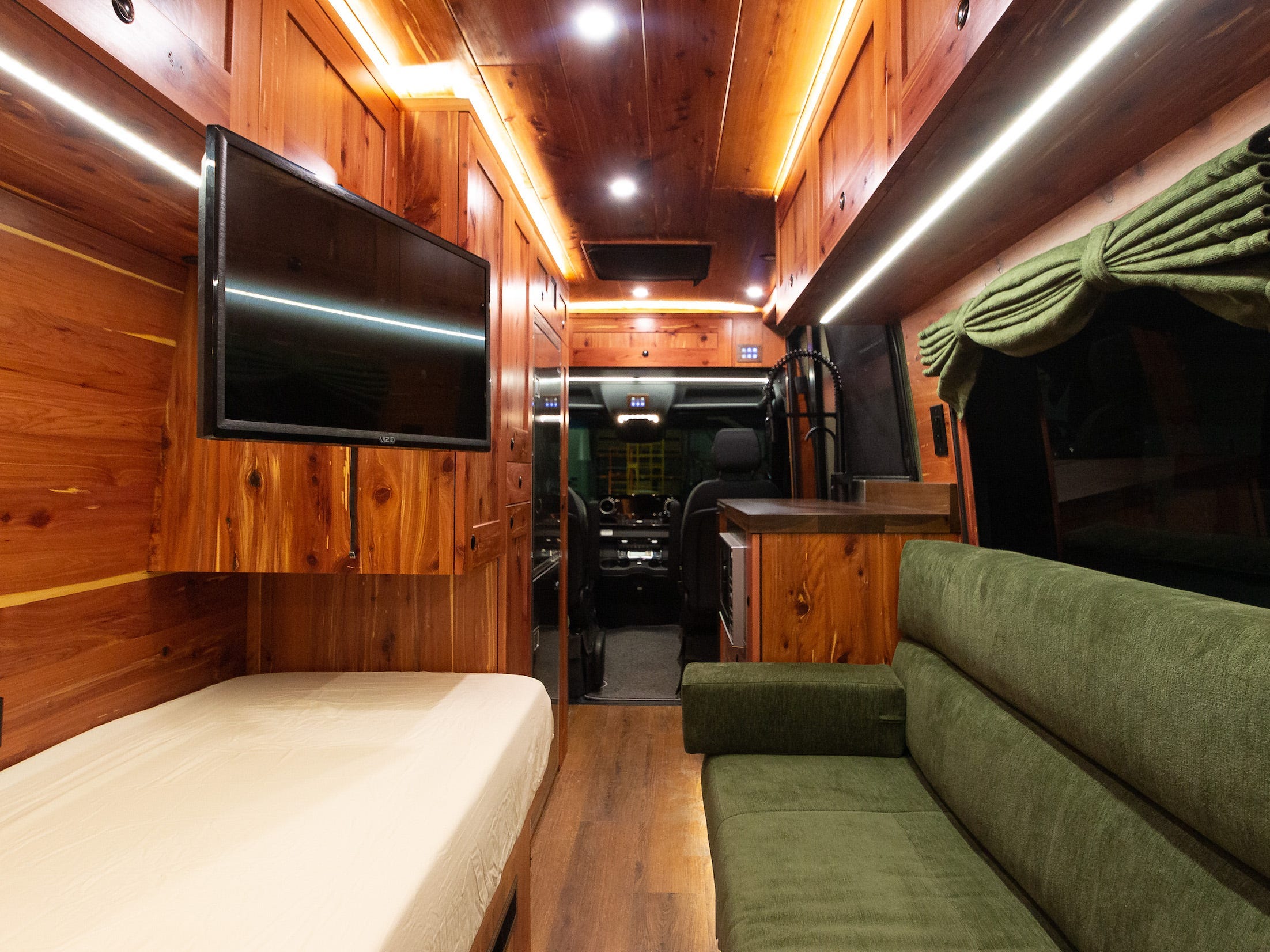 Links ein Bett, rechts eine grüne Couch.  Es gibt einen Fernseher, der von der Decke hängt, und einen Blick durch die Vorderseite des Vans.