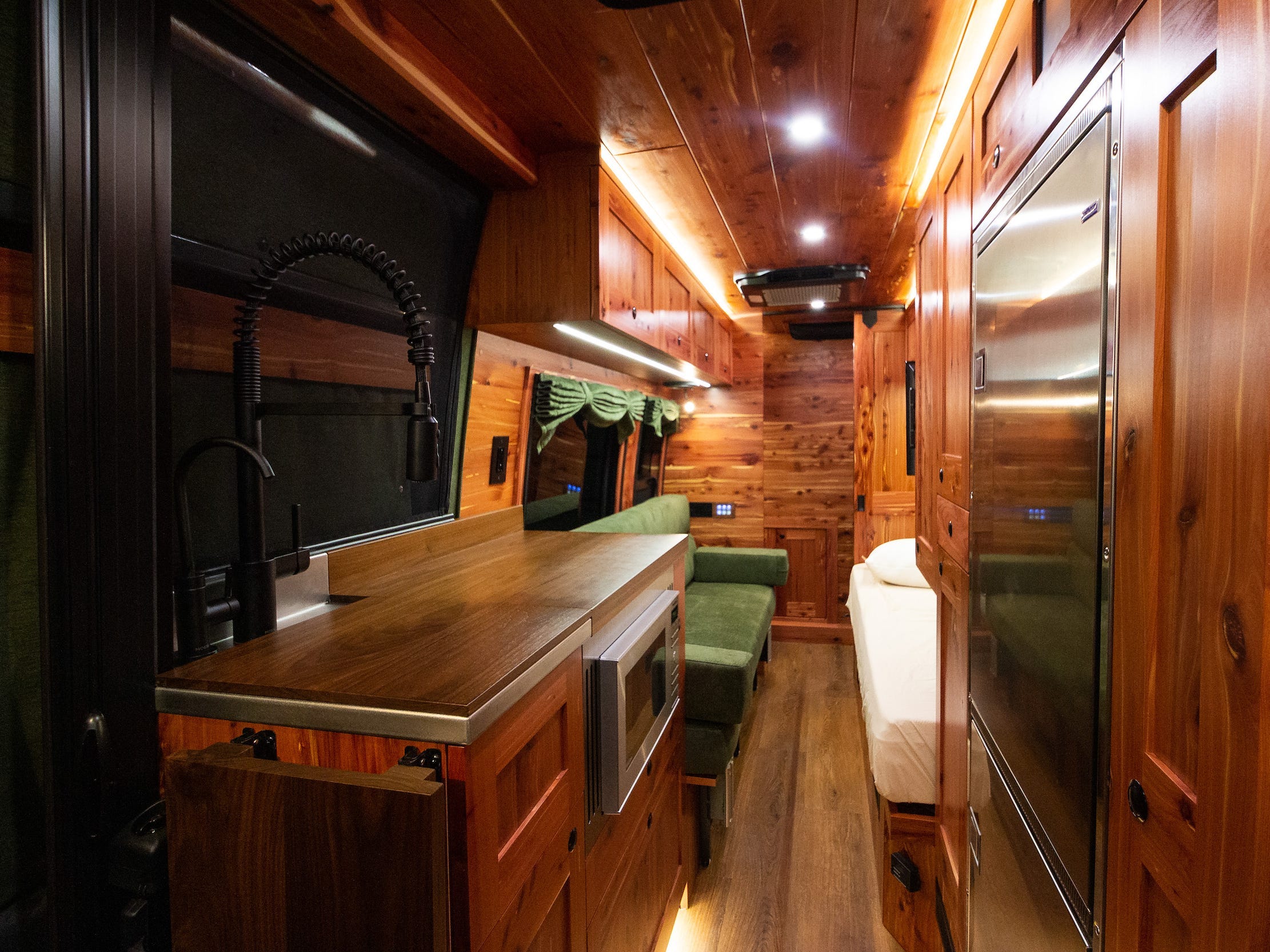 Ein weiter Blick in den Innenraum des Vans zeigt Sitzgelegenheiten, Küche und Bett.  Der Innenraum ist mit dunklem Holz ausgekleidet.