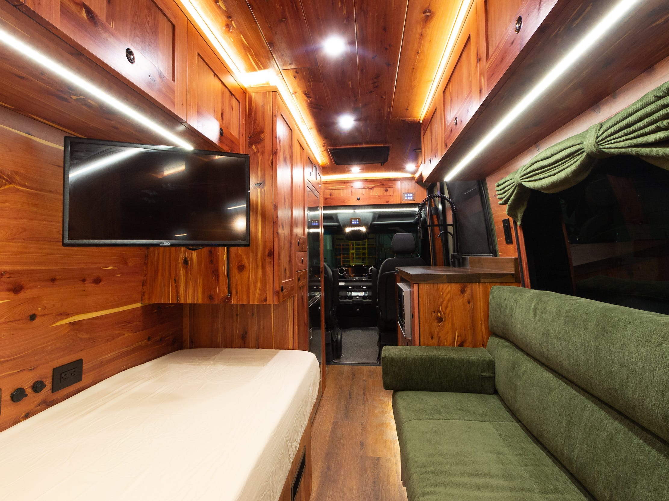 Links ein Bett, rechts eine grüne Couch.  Es gibt einen Fernseher, der von der Decke hängt, und einen Blick durch die Vorderseite des Vans.
