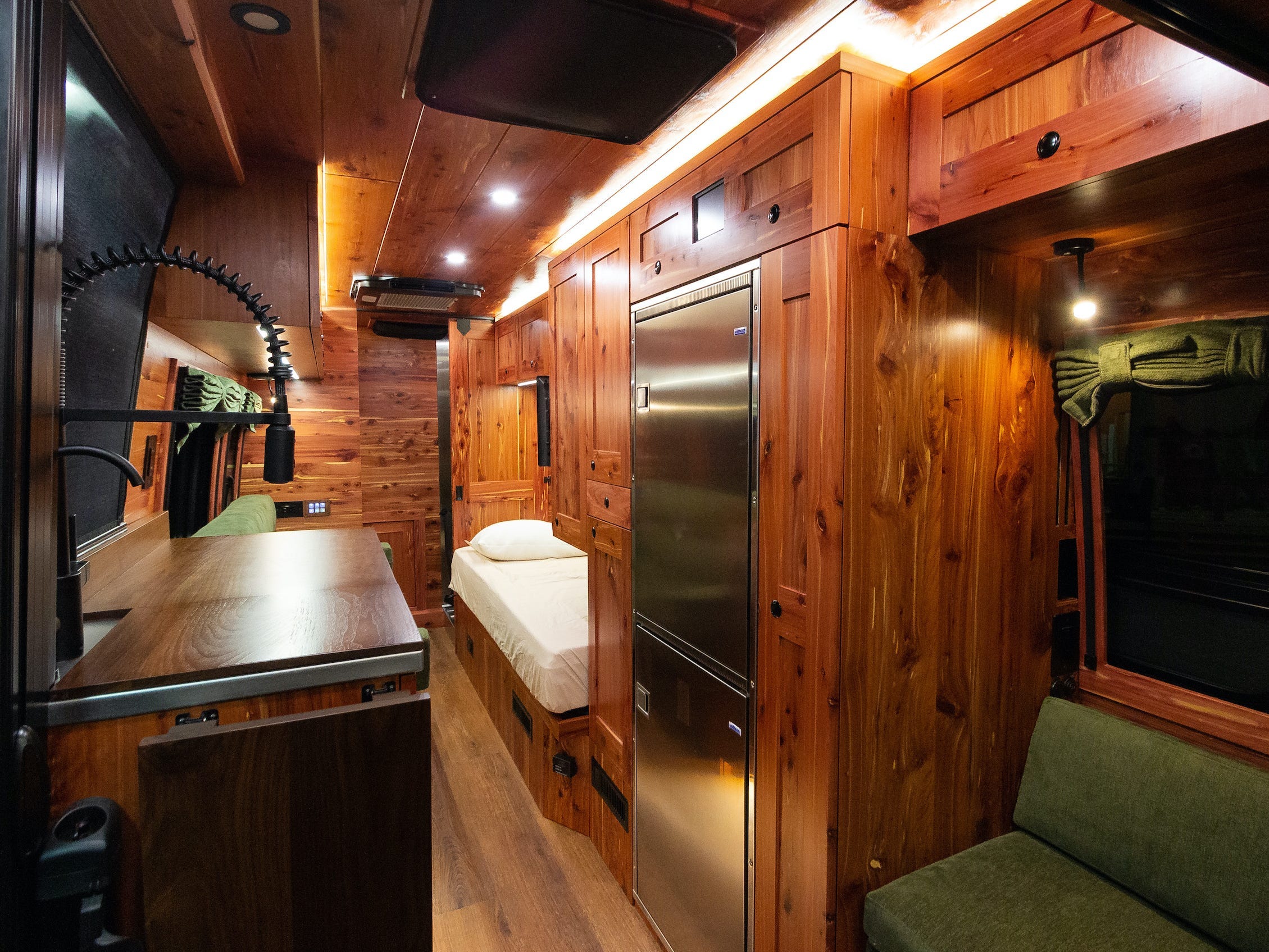 Eine weite Ansicht des Innenraums des Lieferwagens mit Sitz, Küche und Bett.  Der Innenraum ist mit dunklem Holz ausgekleidet.