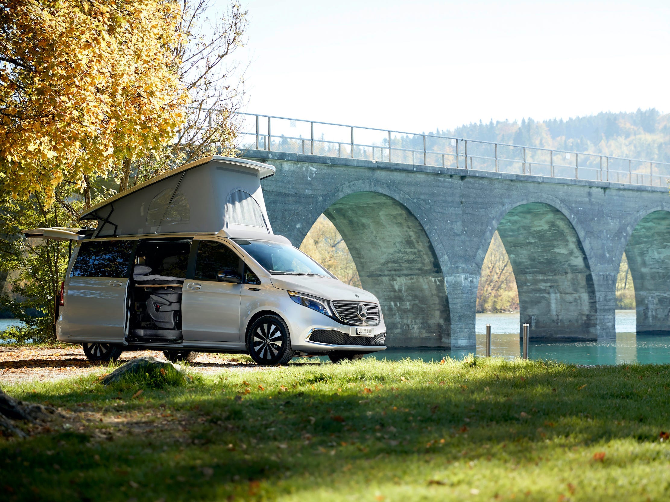 Der Mercedes-Benz EQV mit Aufstelldach und geöffneter Beifahrertür.  Der Van steht neben einem Gewässer und einer Brücke, umgeben von Bäumen, Natur.