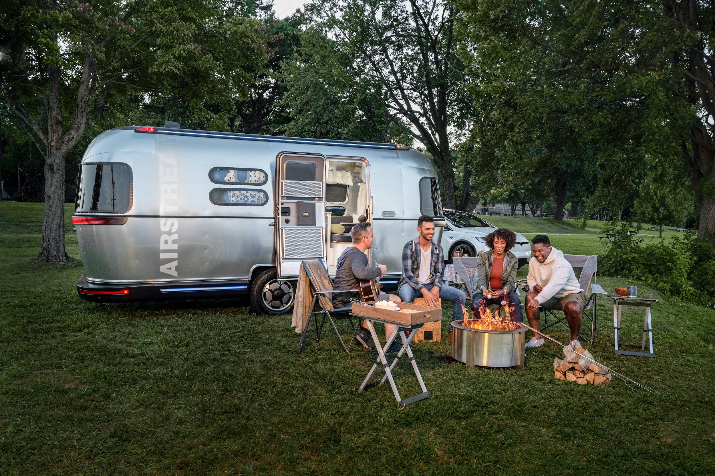 Vier Personen sitzen auf Liegestühlen vor einem Lagerfeuer vor Airstreams Konzept-Wohnwagen.