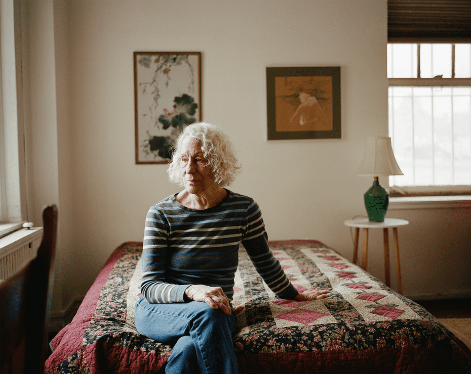 Eine ältere Frau mit weißem lockigem Haar sitzt auf einem Bett mit Steppdecke.