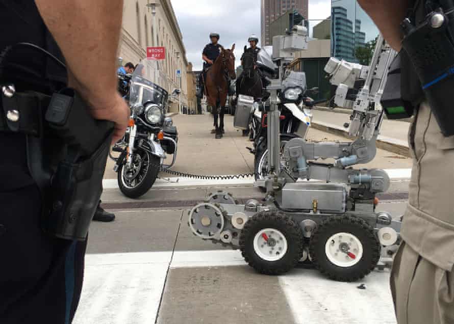 Polizisten stehen in der Nähe eines ferngesteuerten Roboters, während berittene Polizisten im Hintergrund stehen.