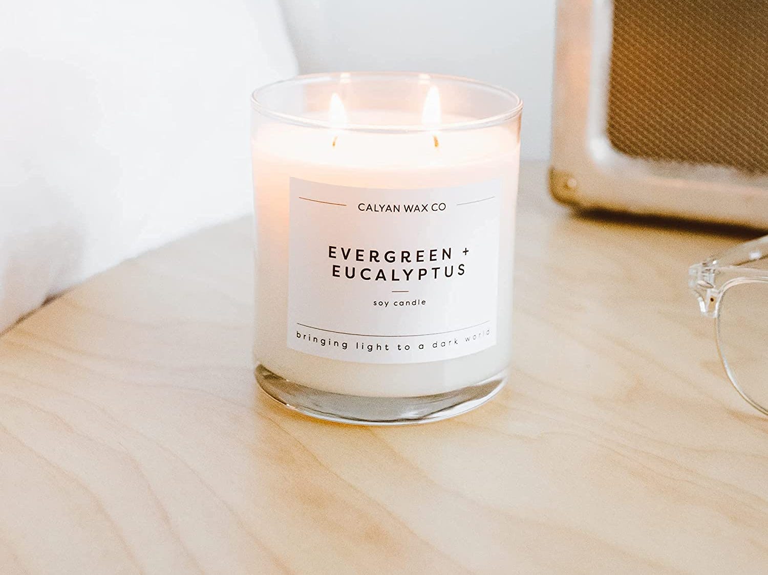 Eine weiß leuchtende Kerze in einem Glasgefäß mit dem Text „Evergreen + Eucalyptus“.  Die Kerze steht auf einem hölzernen Beistelltisch