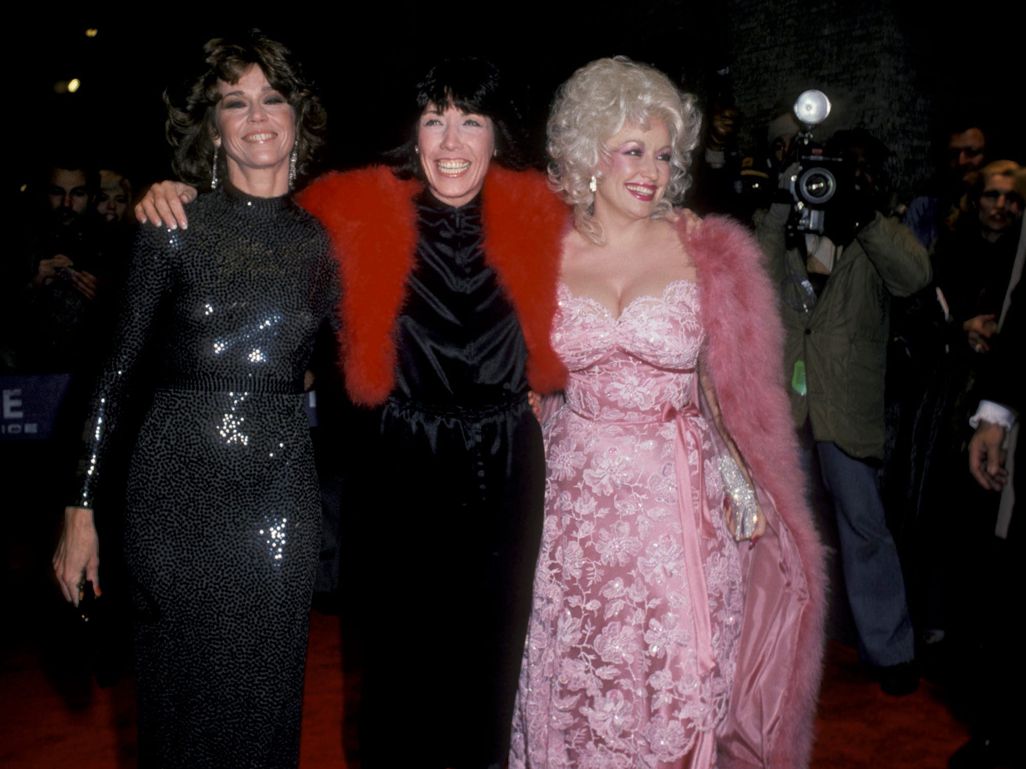 Jane Fonda in einem schwarzen glänzenden Kleid, Lilly Tomlin in einem schwarzen Kleid und einem roten Pelzschal und Dolly Parton in einem babyrosa Blumenspitzenkleid mit einem rosa Pelzmantel.