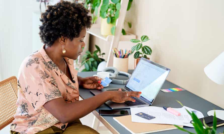 Eine Frau zu Hause in ihrem Heimbüro sitzt an einem Schreibtisch mit einem Laptop beim Online-Shopping.  Sie liest die Details von ihrer Kreditkarte, während sie einen Online-Einkauf tätigt.