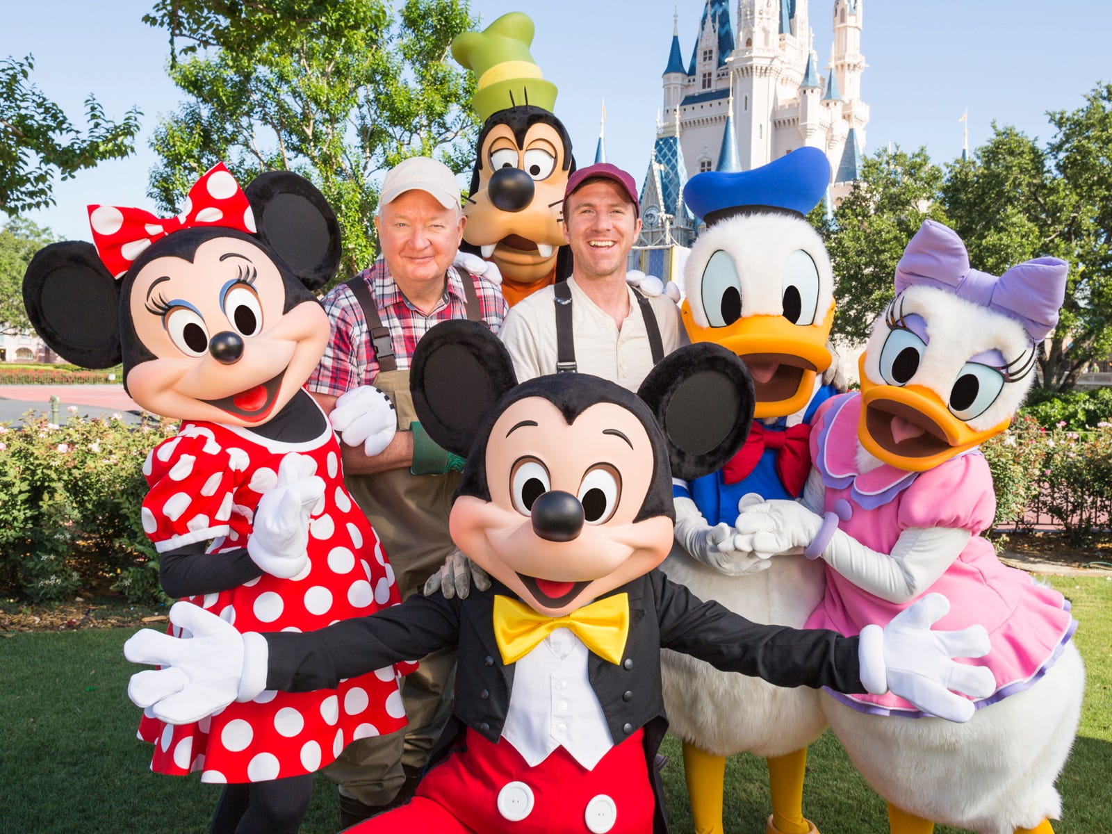 Minnie, Mickey, Goofy, Donald und Daisy Duck und zwei Männer posieren zusammen.