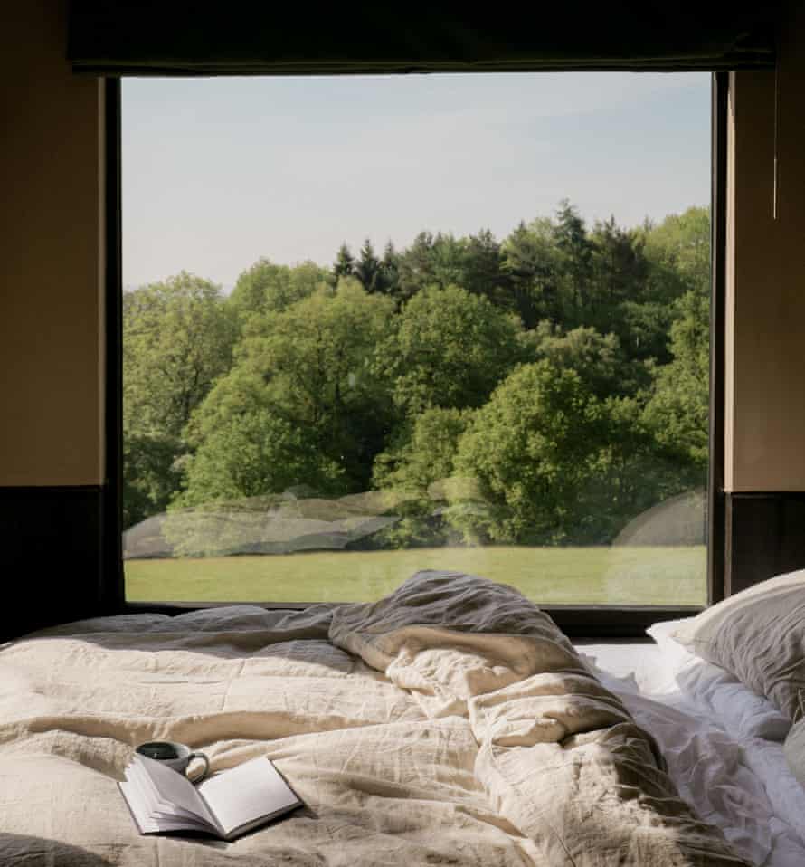 Ein Bett mit einem Buch und einer Tasse Kaffee auf den Laken, mit Blick aus einem großen Fenster auf eine Wiese und Bäume