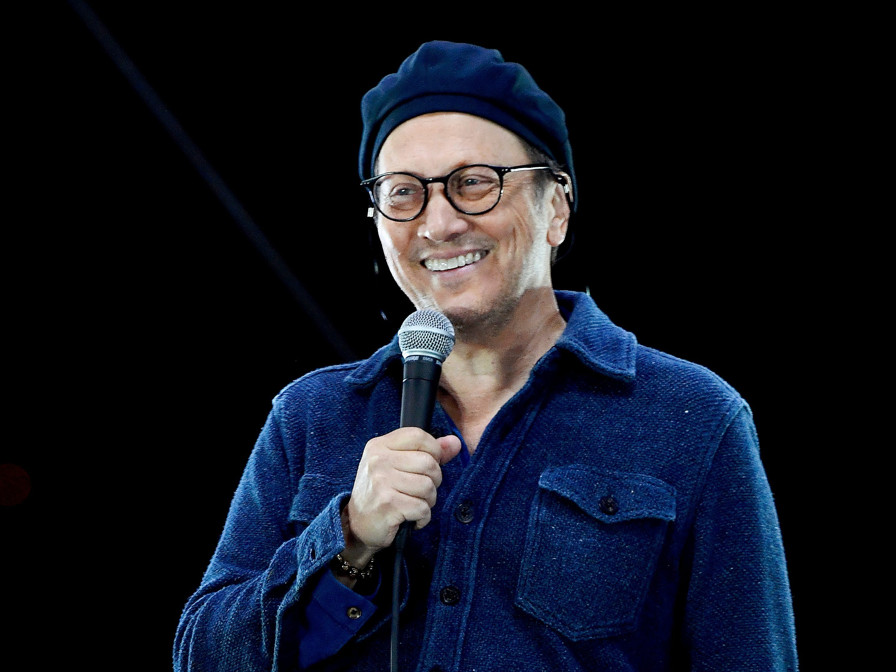 rob schneider trägt ein blaues hemd und einen hut, lächelt auf der bühne und hält ein mikrofon bei der komödie in your cars drive in concert im Jahr 2020