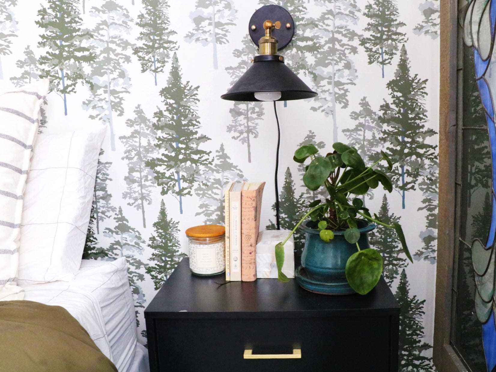Abziehbare Tapete mit weißem und grünem, immergrünem Chasing Paper an der Wand hinter dem Bett mit blauen und weißen Kissen und einem schwarzen Nachttisch mit Büchern, einer Pflanze und einer Wandleuchte aus schwarzem Metall darüber.