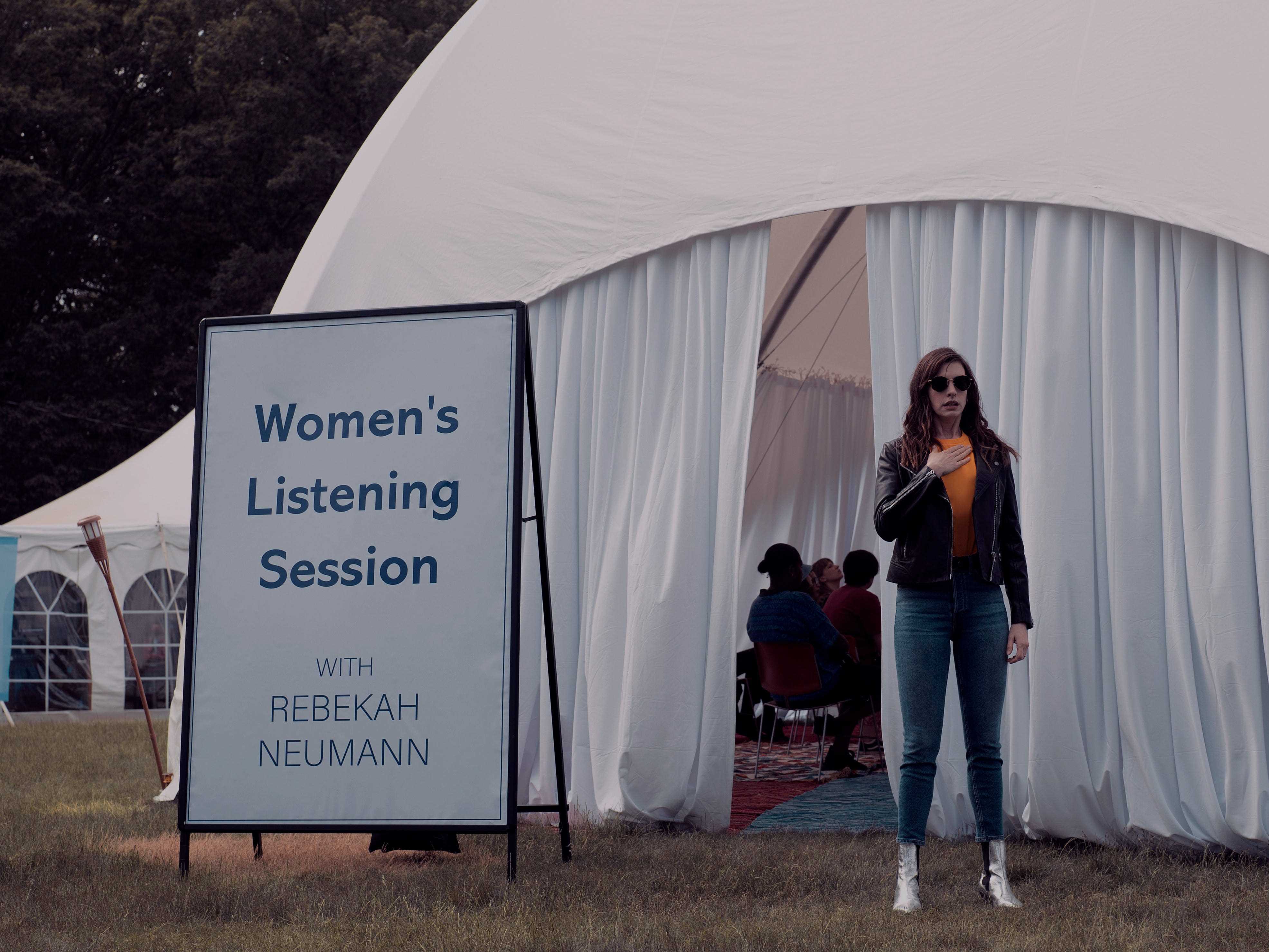 Anne Hathaway als Rebekah Neumann steht vor einem Zelt, an dem ein Schild mit der Aufschrift „Hörseminar für Frauen“ angebracht ist