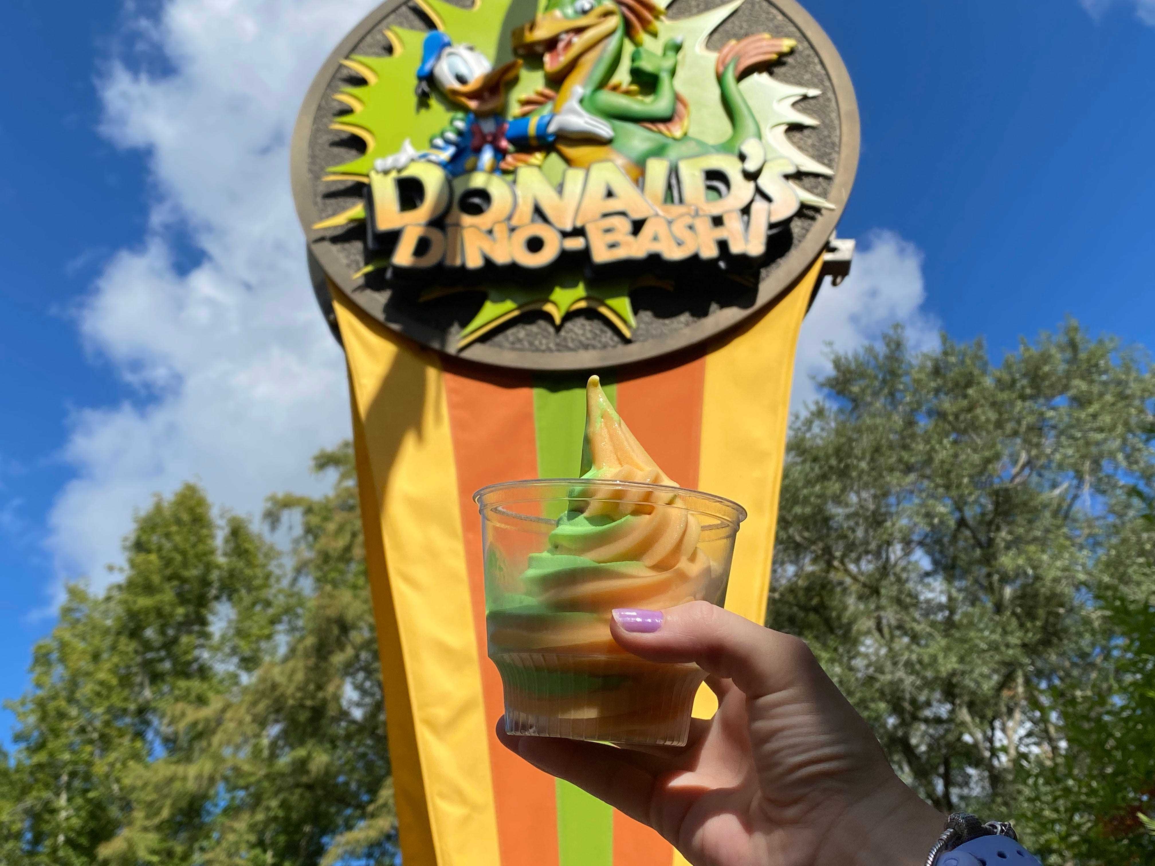 Hand, die eine Tasse mit grünem und orangefarbenem Softeis im Disney-Tierreich hält