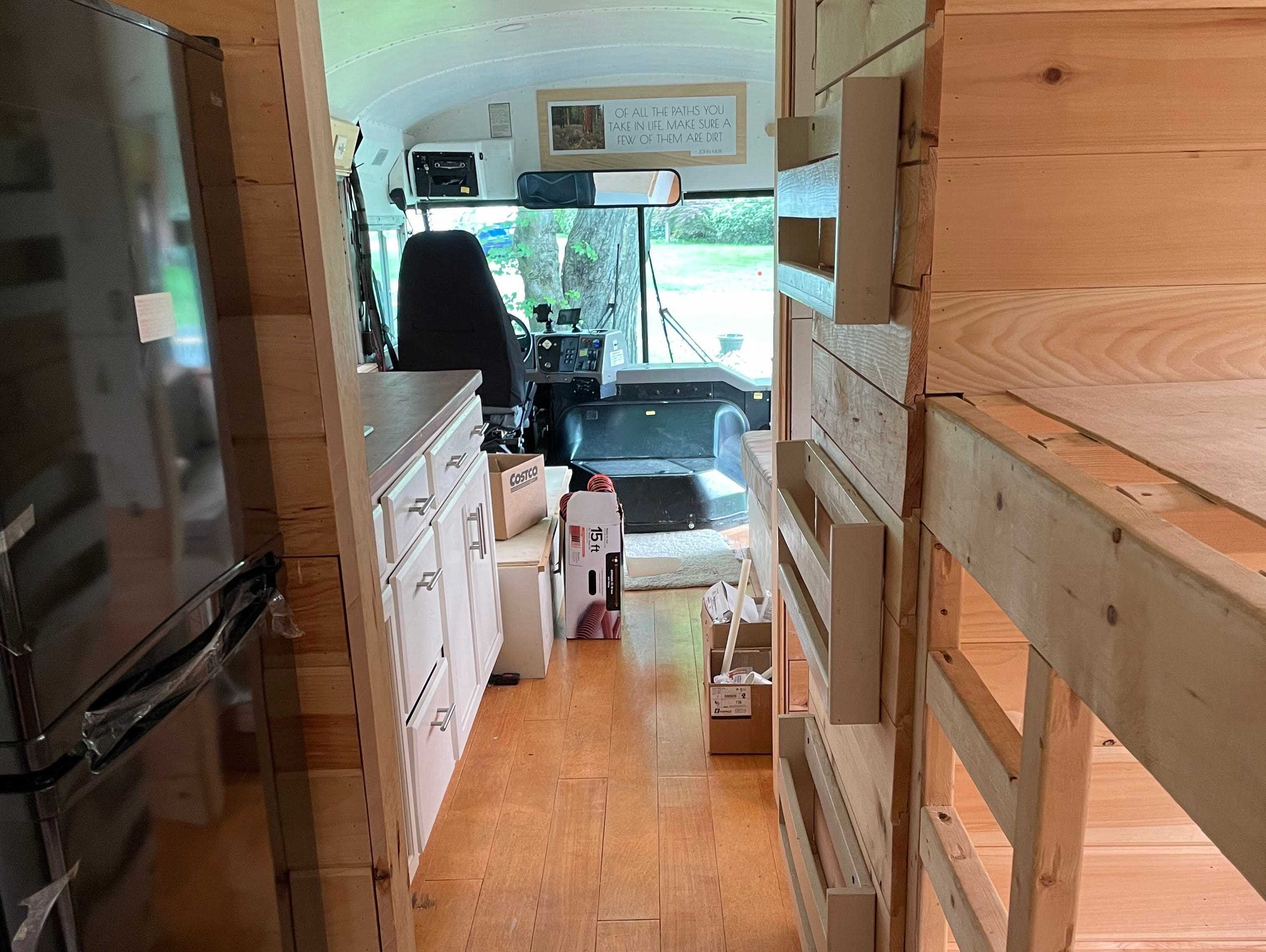 Holzregale und Schubladen und schwarzer Kühlschrank mit Blick auf weiße Schränke und Fahrerbereich vor dem neu gestalteten Schulbus