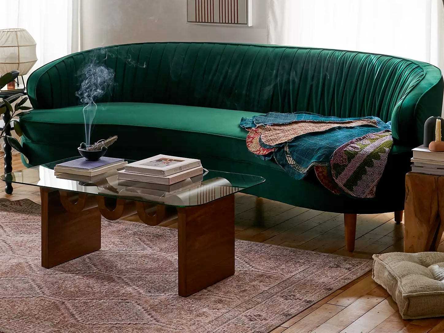 Ein Camila-Sofa von Urban Outfitters aus grünem Samt steht in einem Wohnzimmer vor einem Fenster neben einem Couchtisch.