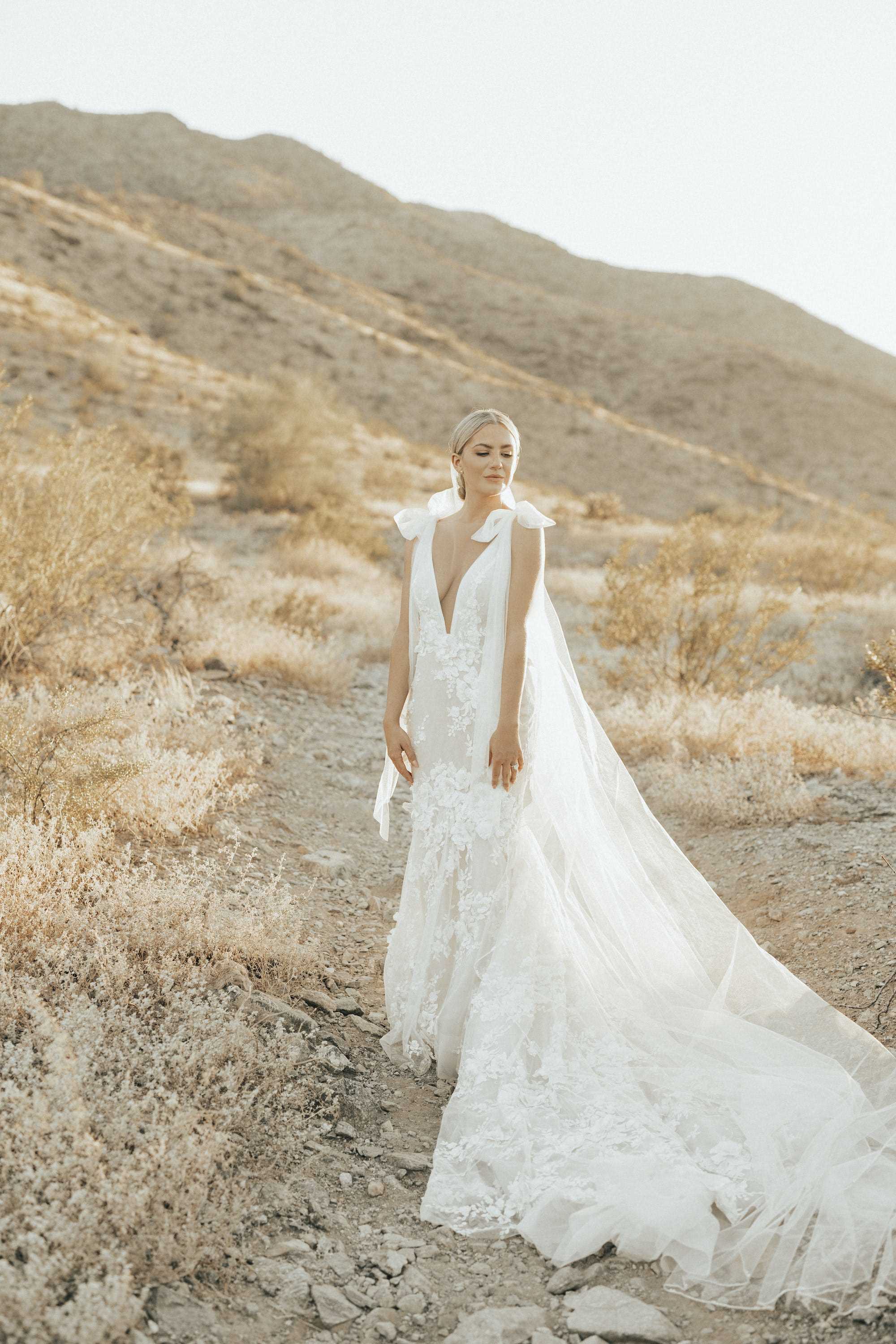 Eine Braut blickt in ihrem Hochzeitskleid vor hügeligem Hintergrund in die Ferne.