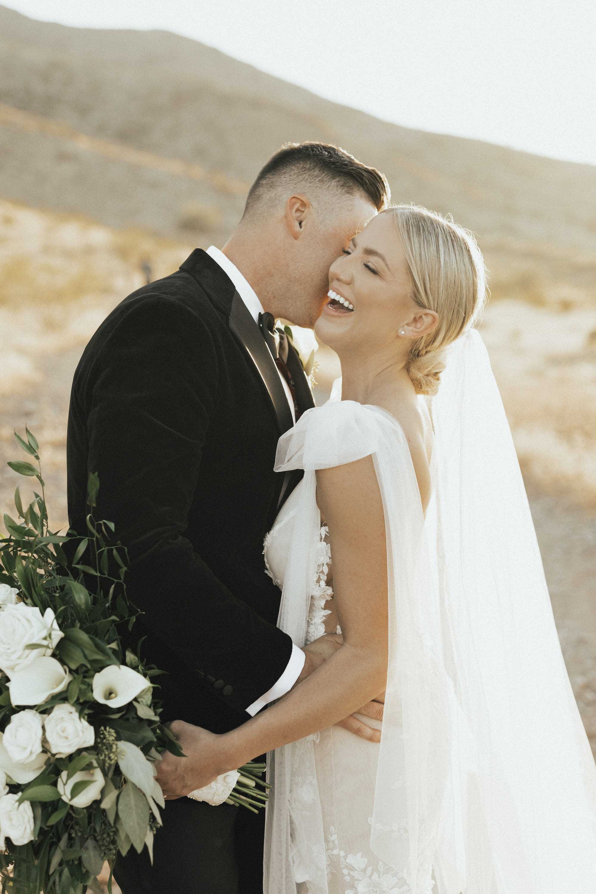 Eine Braut lacht und hält einen Blumenstrauß, während ihr Bräutigam sie auf die Wange küsst.