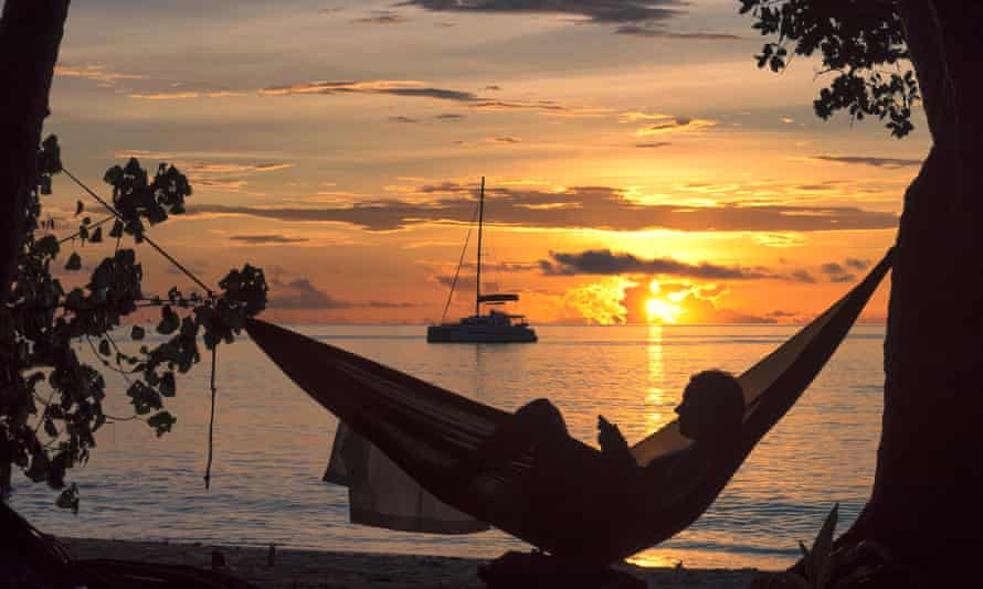 Silhouette einer Frau, die bei Sonnenuntergang in der Hängematte vor einer Yacht in einer tropischen Bucht liest J53Y8F Strandurlaub, Silhouette einer Frau, die bei Sonnenuntergang auf einer tropischen Insel in der Hängematte liest.