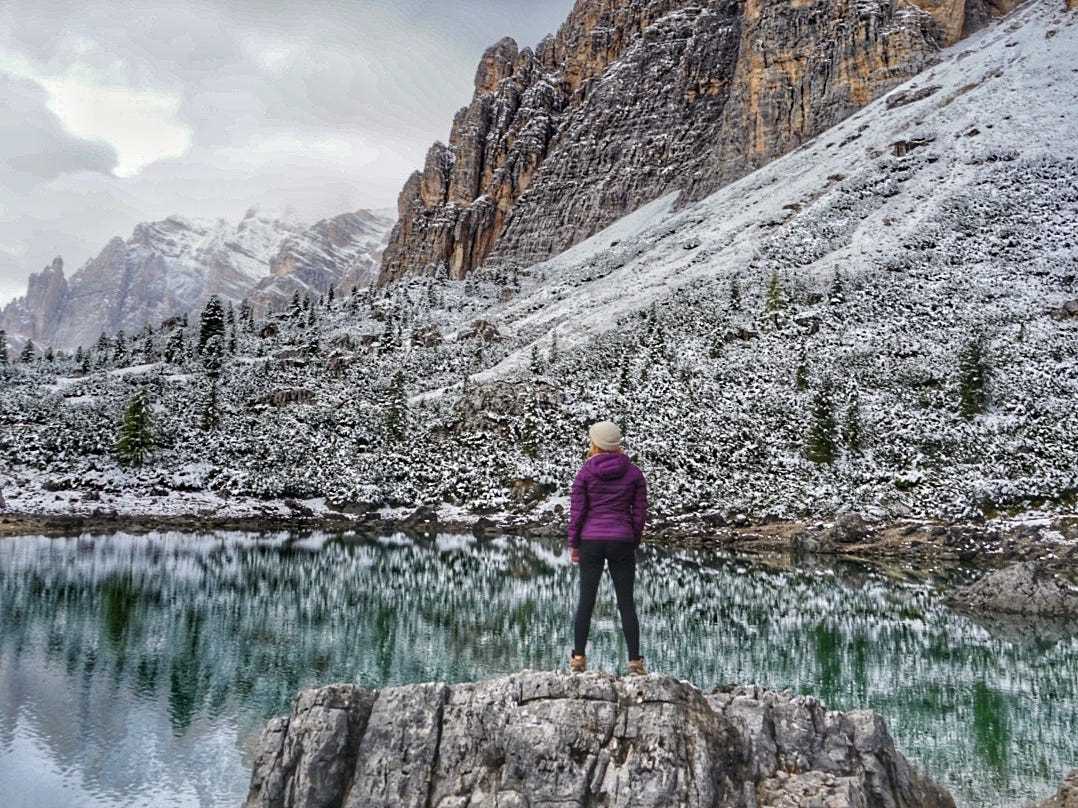 Eine Frau, die auf einem Felsen inmitten eines Sees steht, umgeben von Bergen und Schnee.