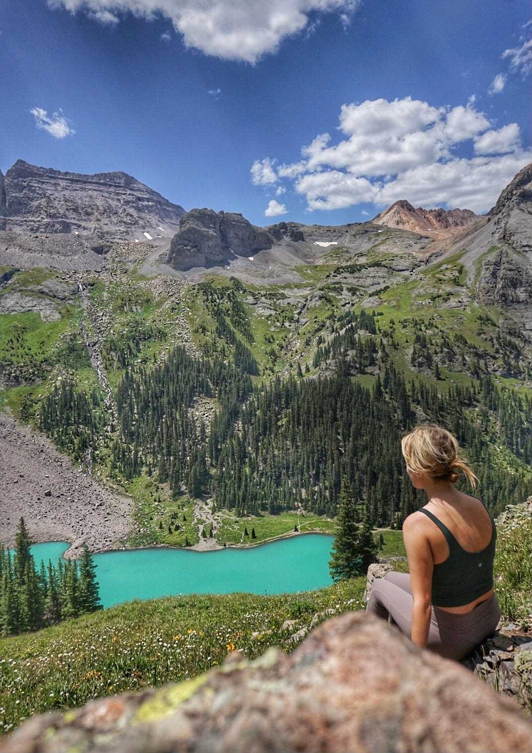 Eine Frau mit dem Rücken zur Kamera mit Blick auf einen blauen See, Berge und Bäume.