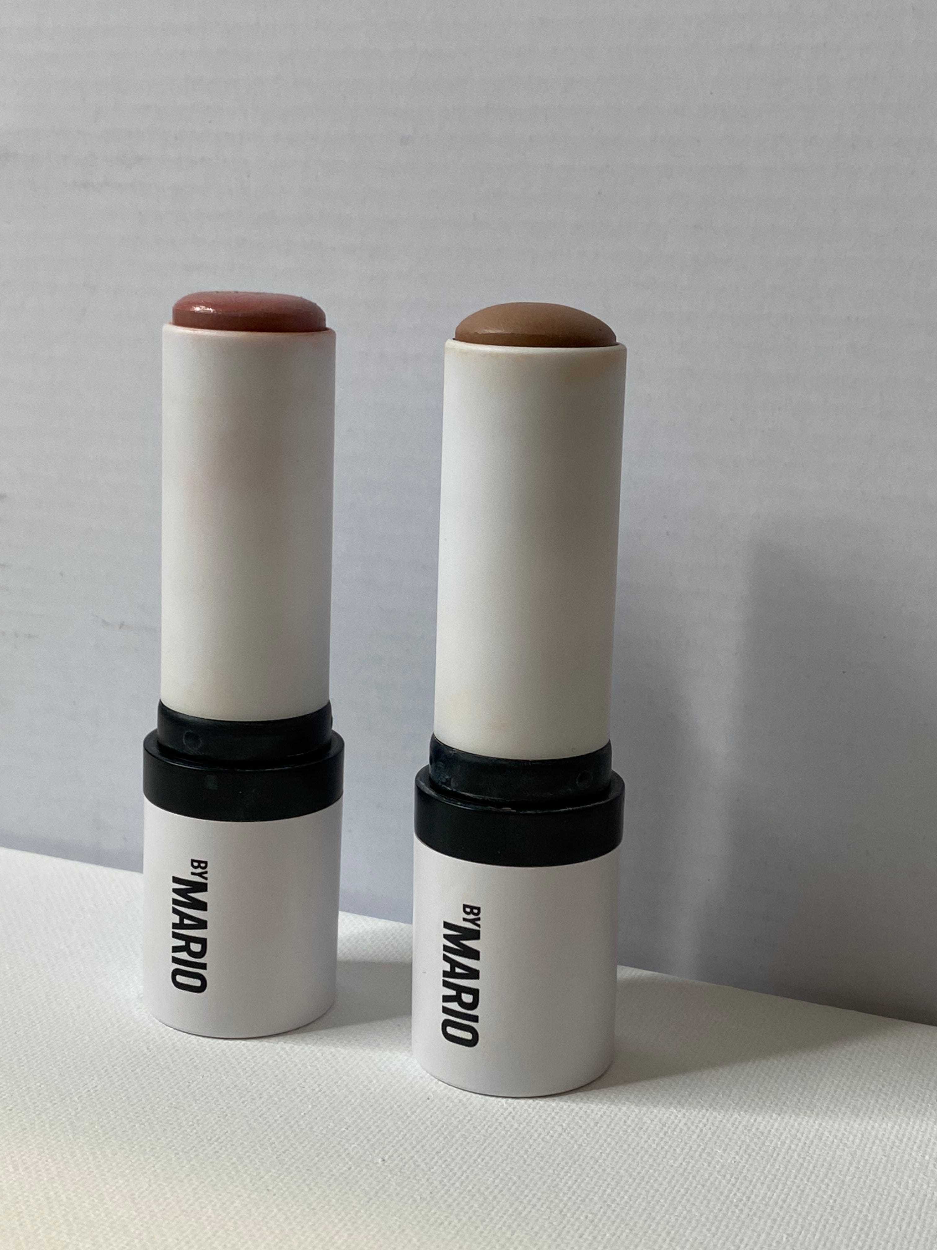 Zwei cremefarbene Make-up-Sticks mit dem Schriftzug „by MARIO“ auf der Seite.  Das Produkt auf der linken Seite ist rosa und das Produkt auf der rechten Seite ist braun.