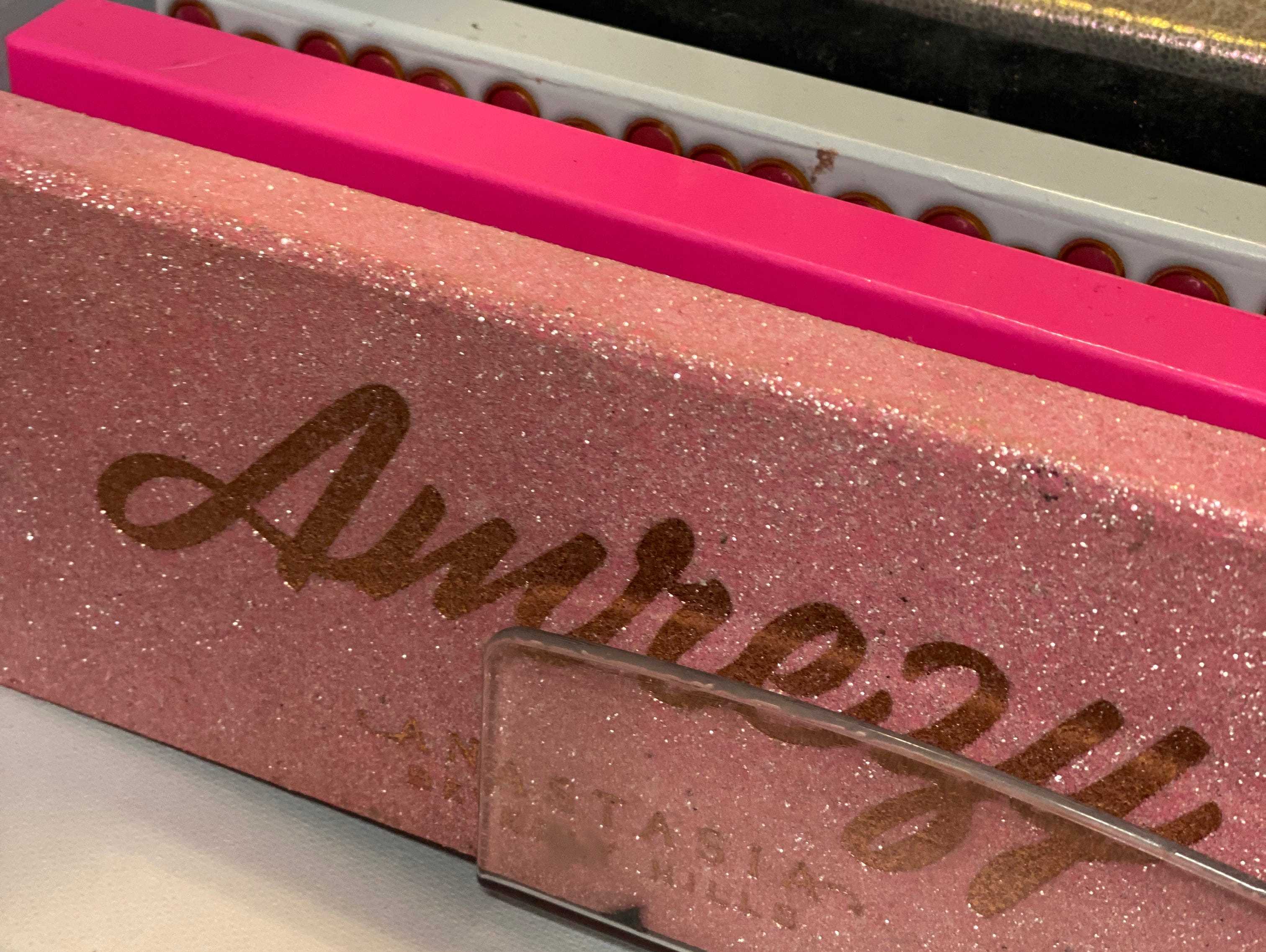 Sammlung von Lidschattenpaletten in einer Schublade, bei der außer der vorderen Palette nur Stacheln sichtbar sind.  Die vordere Palette ist rosa und glitzert mit goldenem Text, auf dem „Amrezy“ in großen Buchstaben und kleiner „Anastasia Beverly Hills“-Text darunter steht