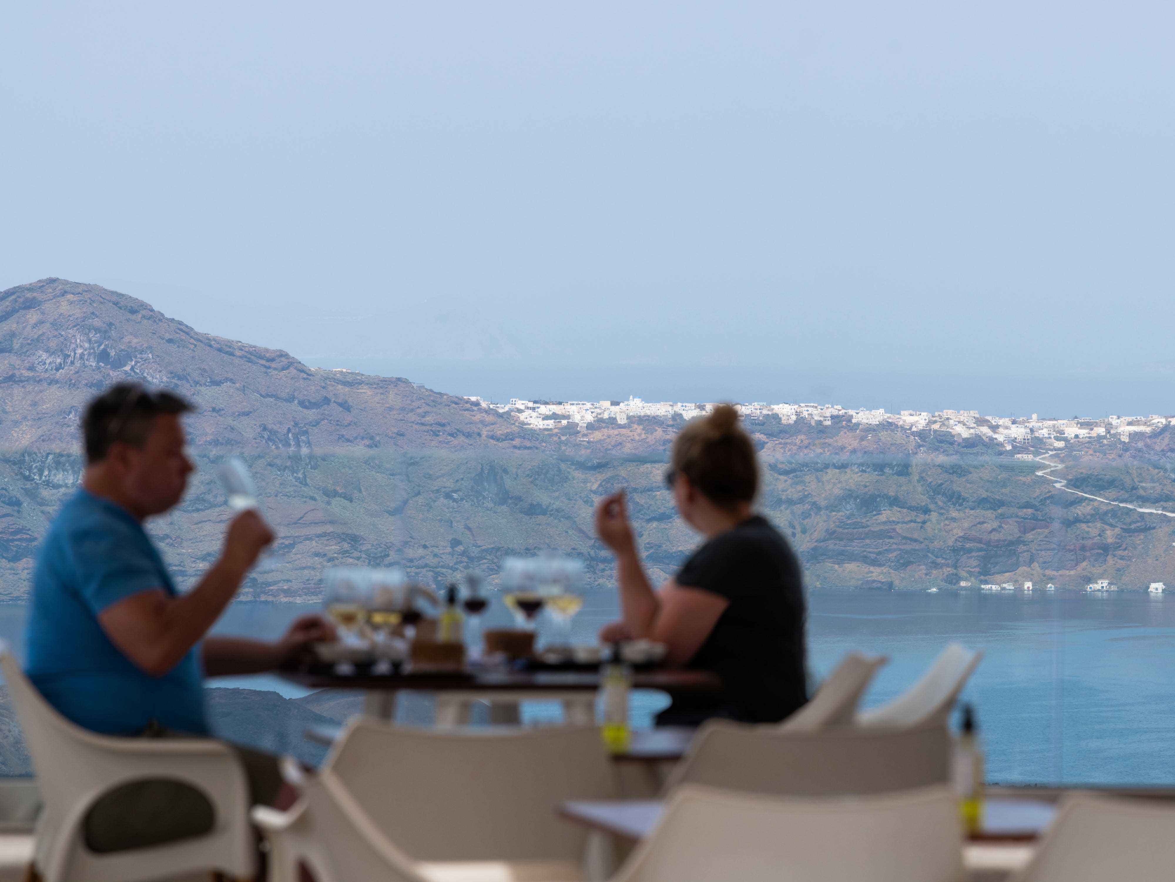 Paar sitzt am Tisch beim Essen mit Blick auf die Küste und das Meer mit weißen Stühlen im Vordergrund
